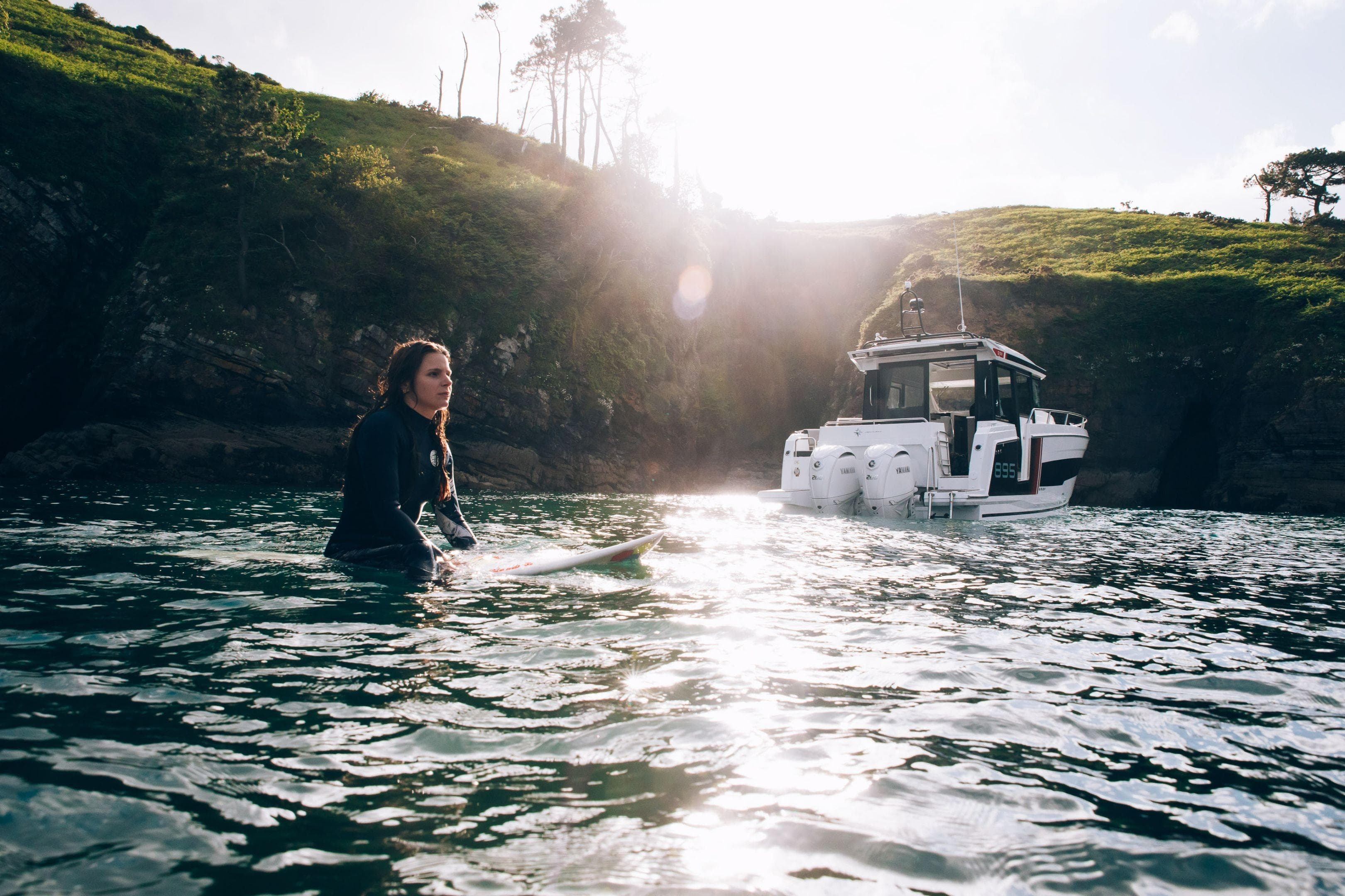 Surfer near Jeanneau NC 895 Sport Series 2 boat, 2026 model, in scenic coastal waters.