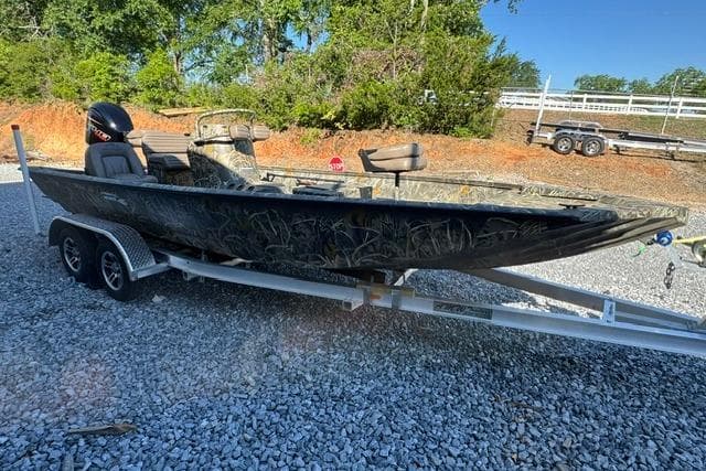 2026 War Eagle 2170 Blackhawk boat on trailer, parked on gravel, surrounded by trees.