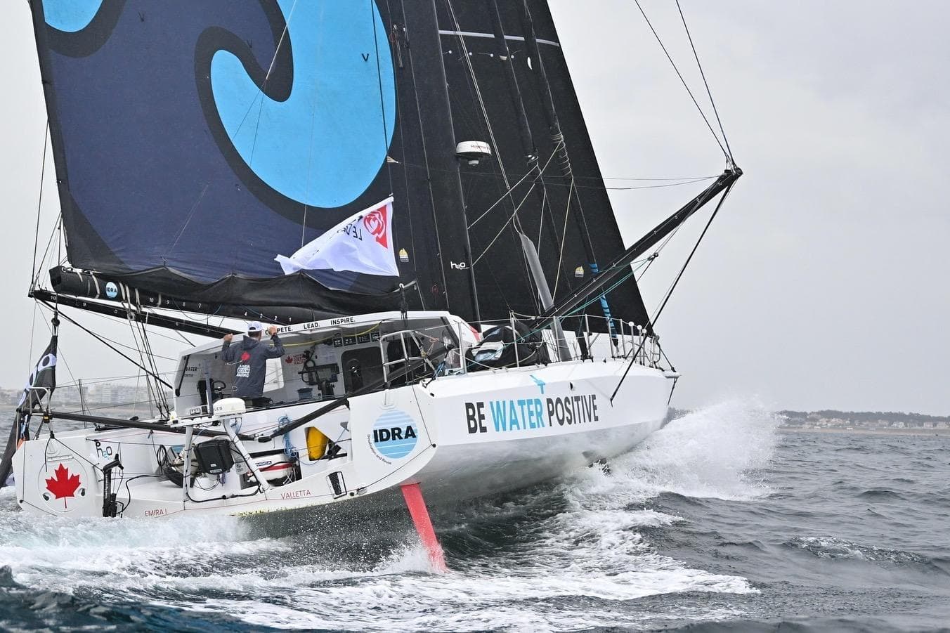 Sailing yacht IMOCA 60 navigating waves, displaying "Be Water Positive" slogan.