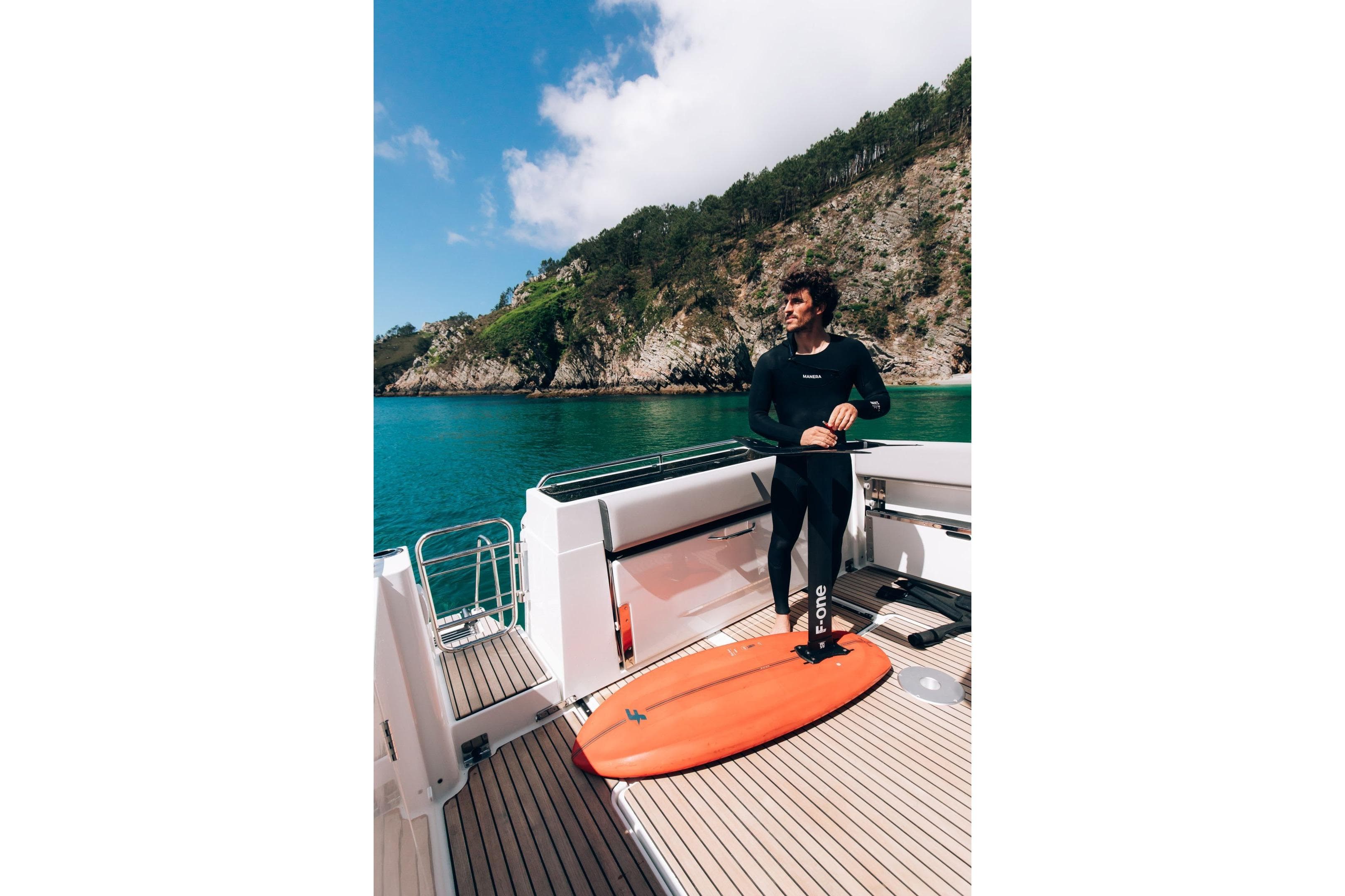 Man on Jeanneau NC 895 Sport Series 2 boat with surfboard, scenic coastal backdrop.