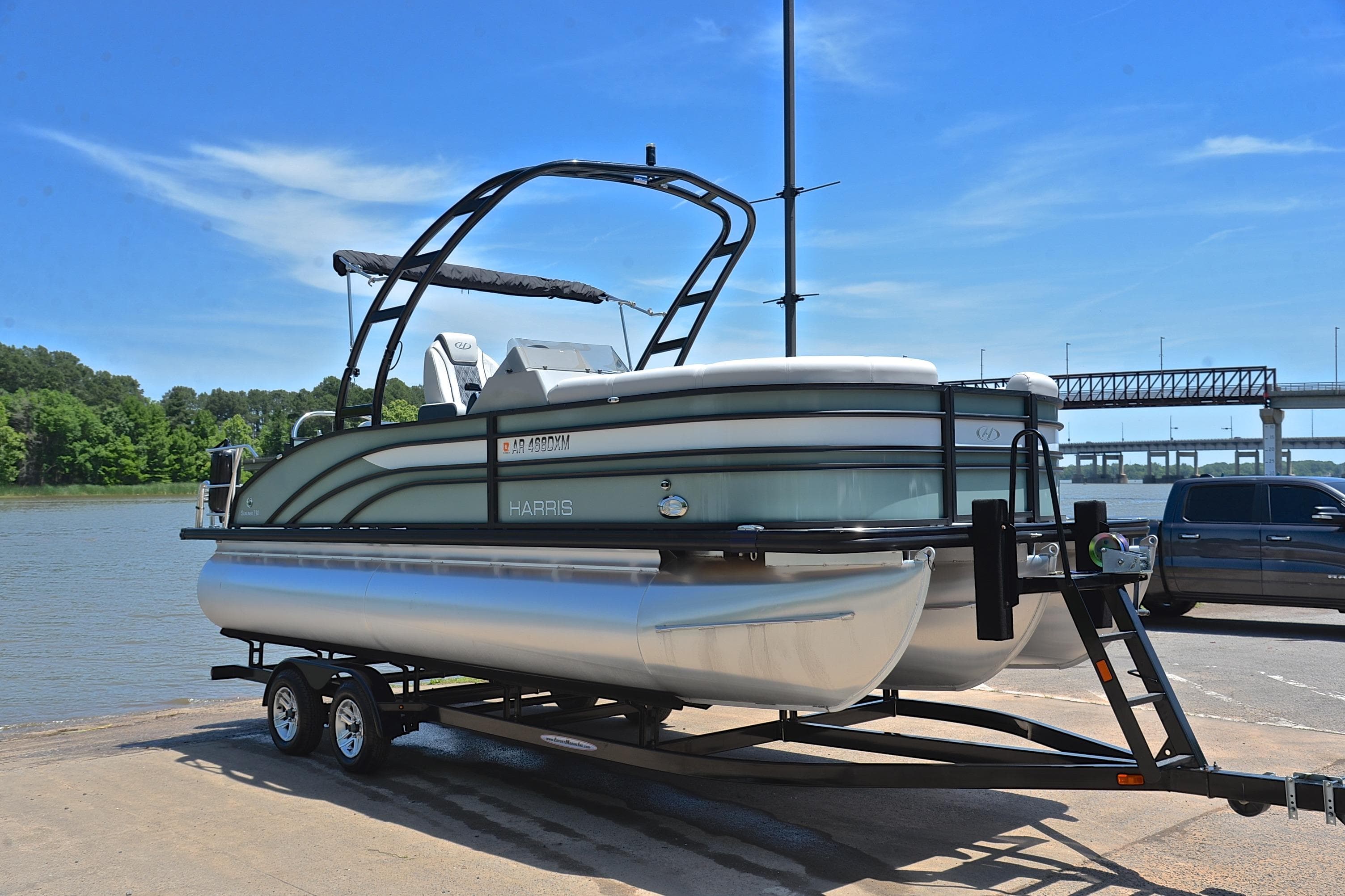 2023 Harris Sunliner 230 pontoon boat on trailer by riverside, clear sky background.