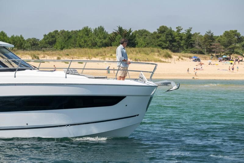 2025 Jeanneau NC 1295 Coupe yacht near a sandy beach with trees in the background.
