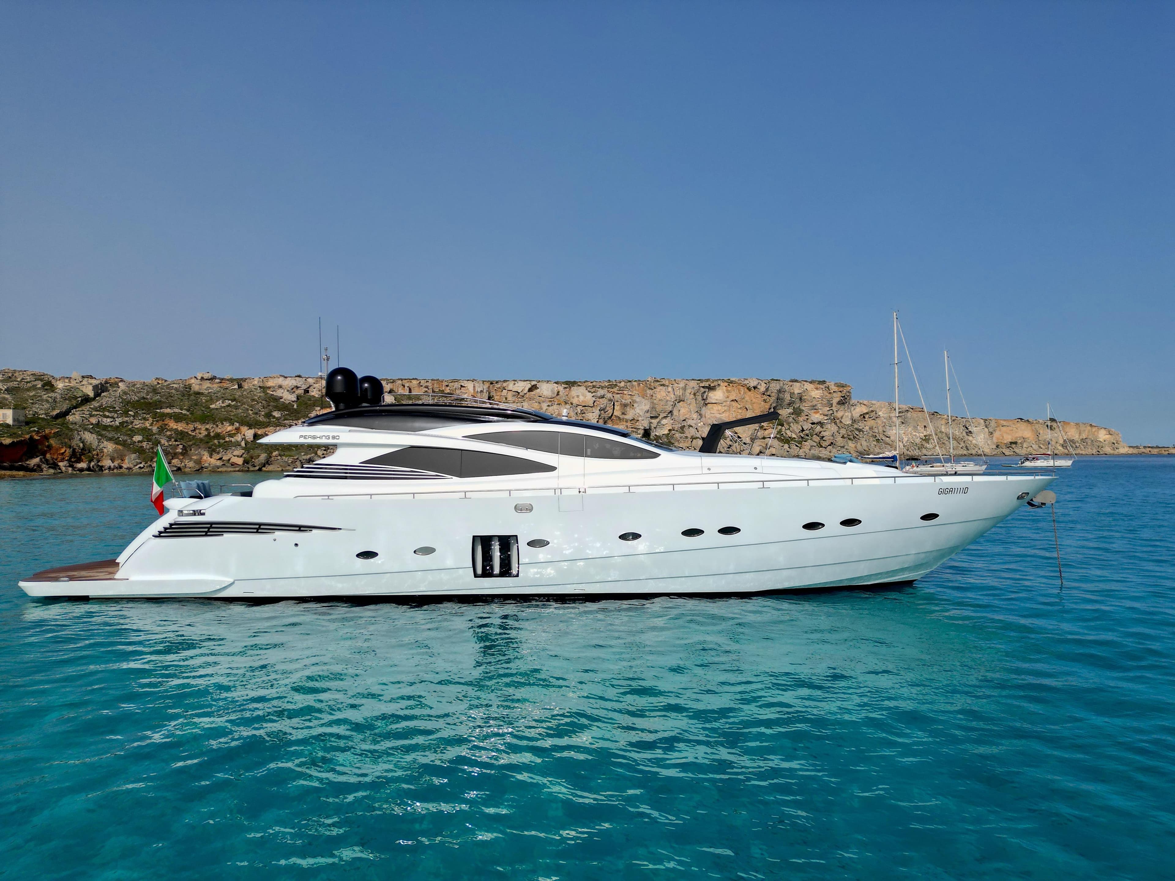 2008 Pershing 90 yacht on clear blue water with rocky coastline in the background.