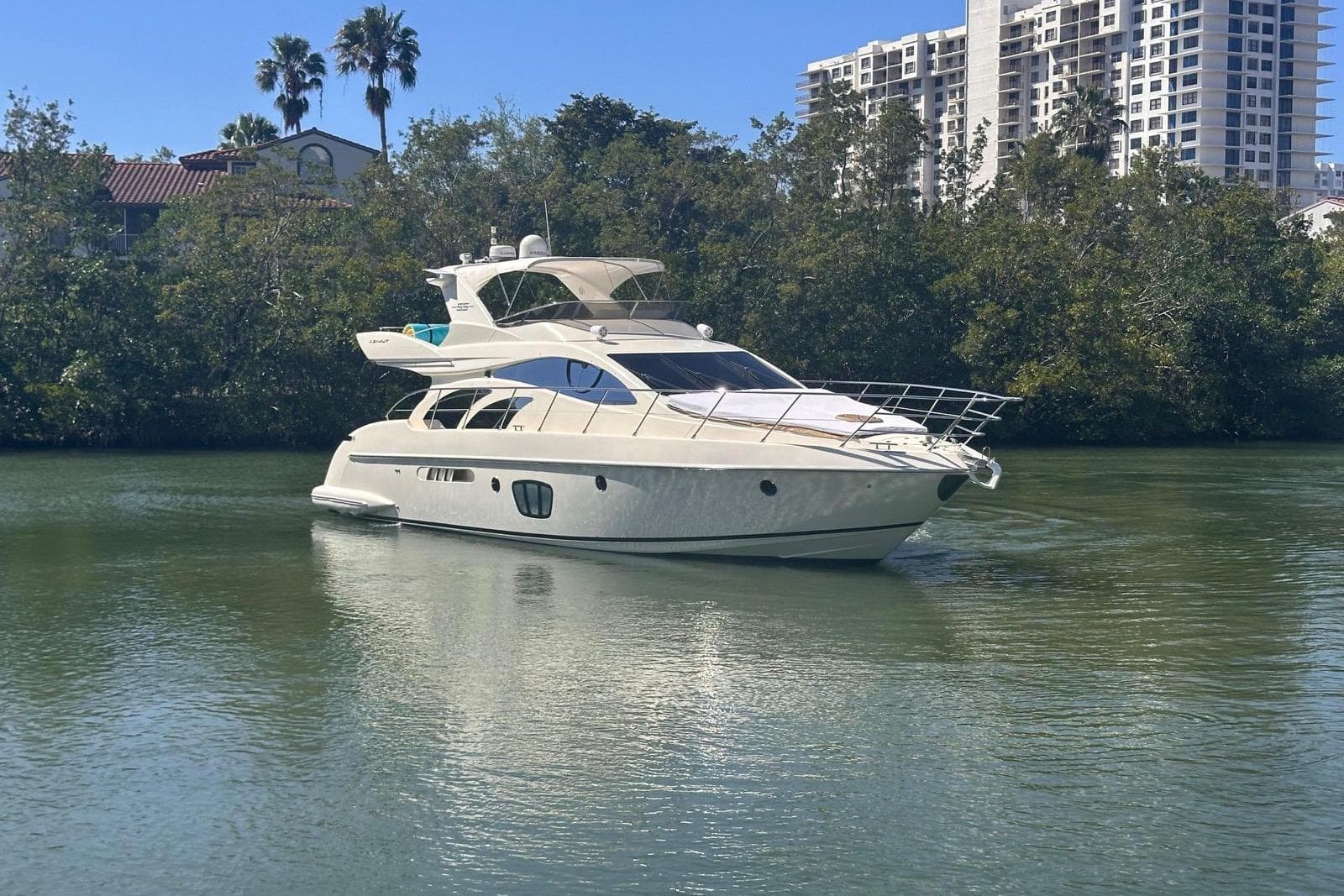 2007 Azimut 55E yacht cruising on a calm river with lush greenery and buildings in the background.
