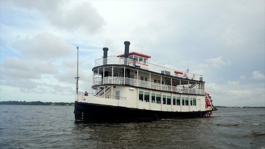 Vintage 1984 custom paddle wheel boat cruising on a calm river under cloudy skies.