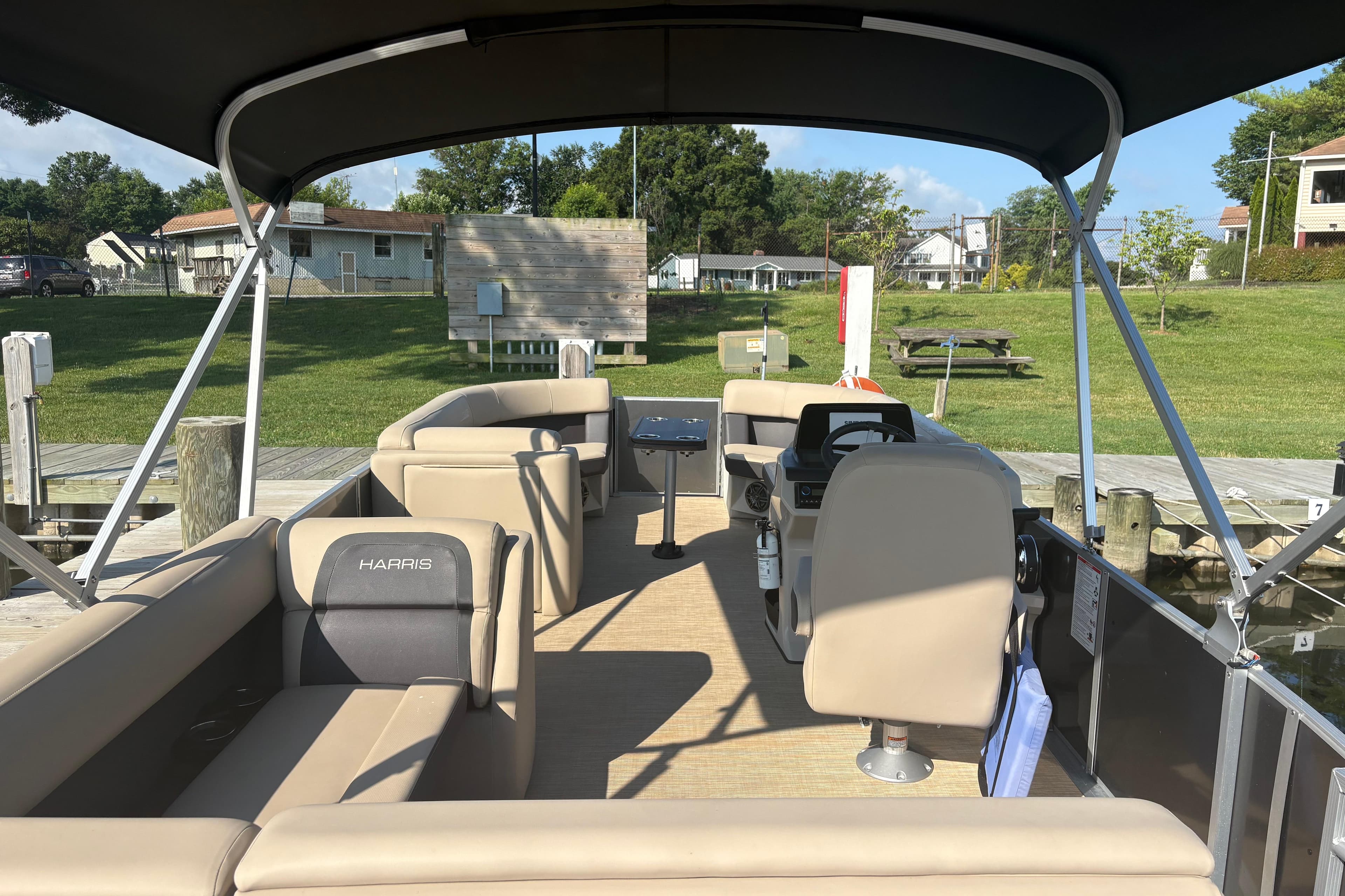 2024 Harris HCX23 pontoon boat interior with seating and helm, docked by grassy area.