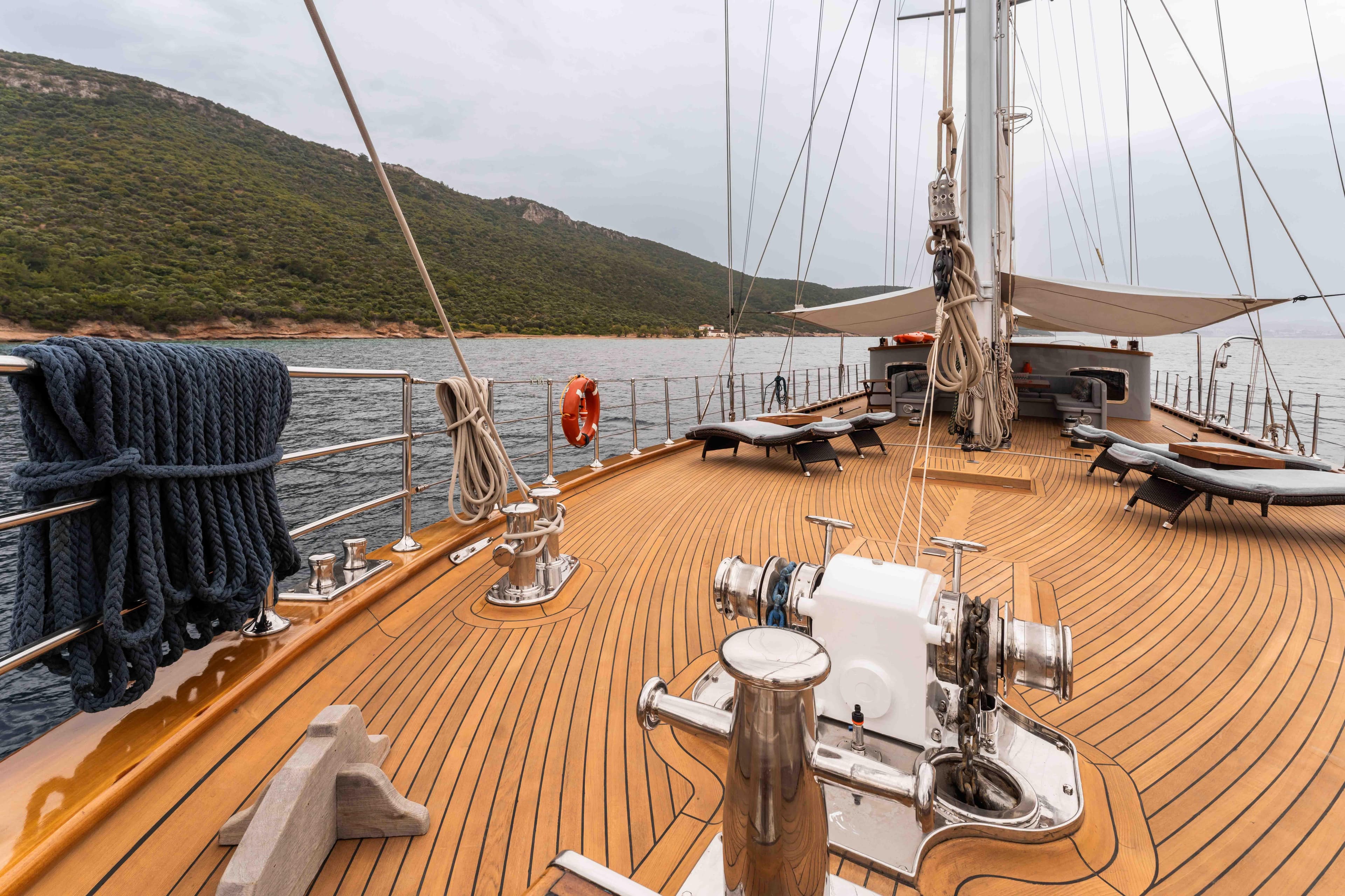Custom 2009 sailing yacht with wooden deck, anchored near a lush green coastline.