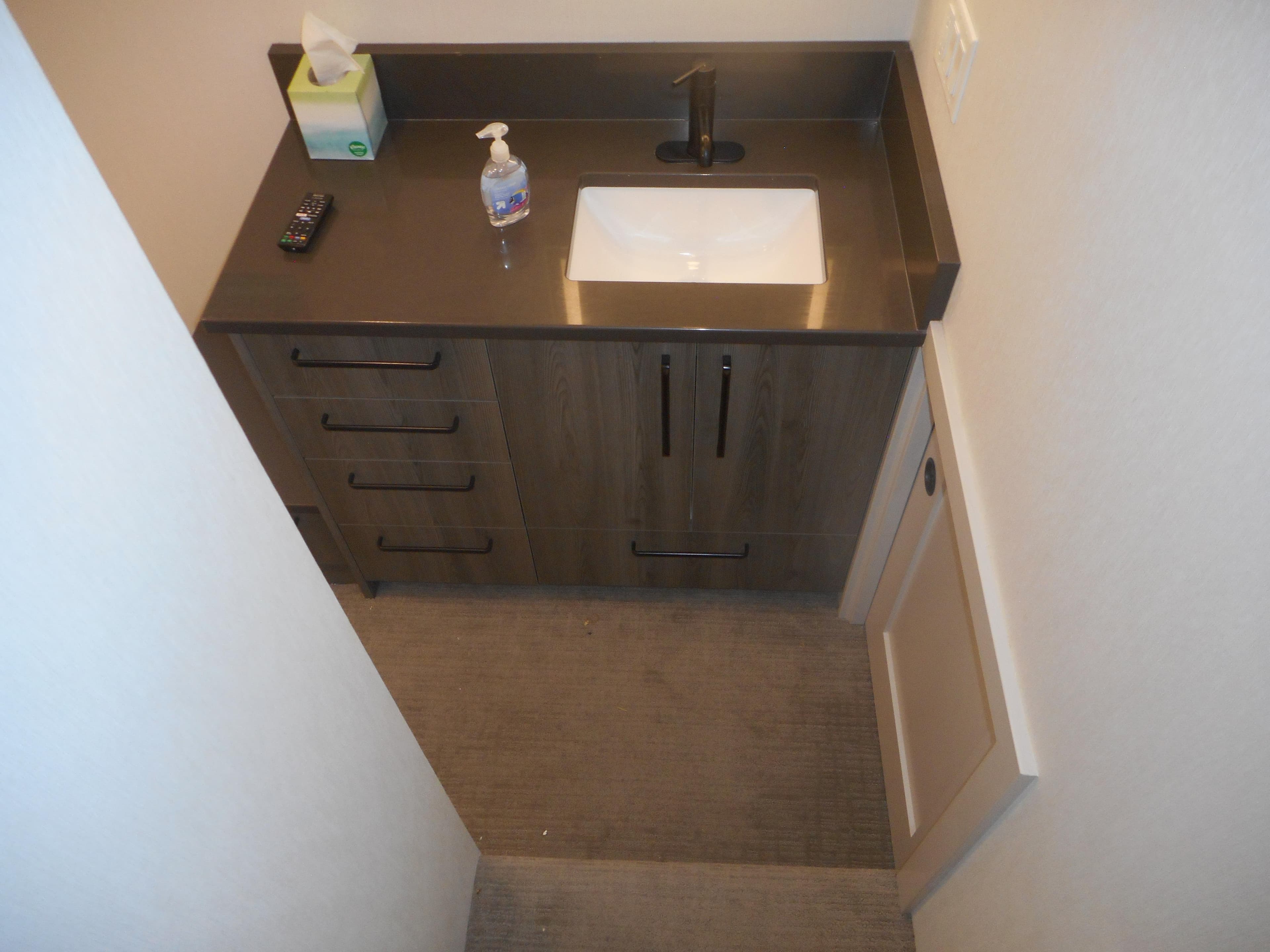 Compact bathroom vanity with dark countertop, sink, and storage drawers in Adonia Obsession Trip #12, 2021.