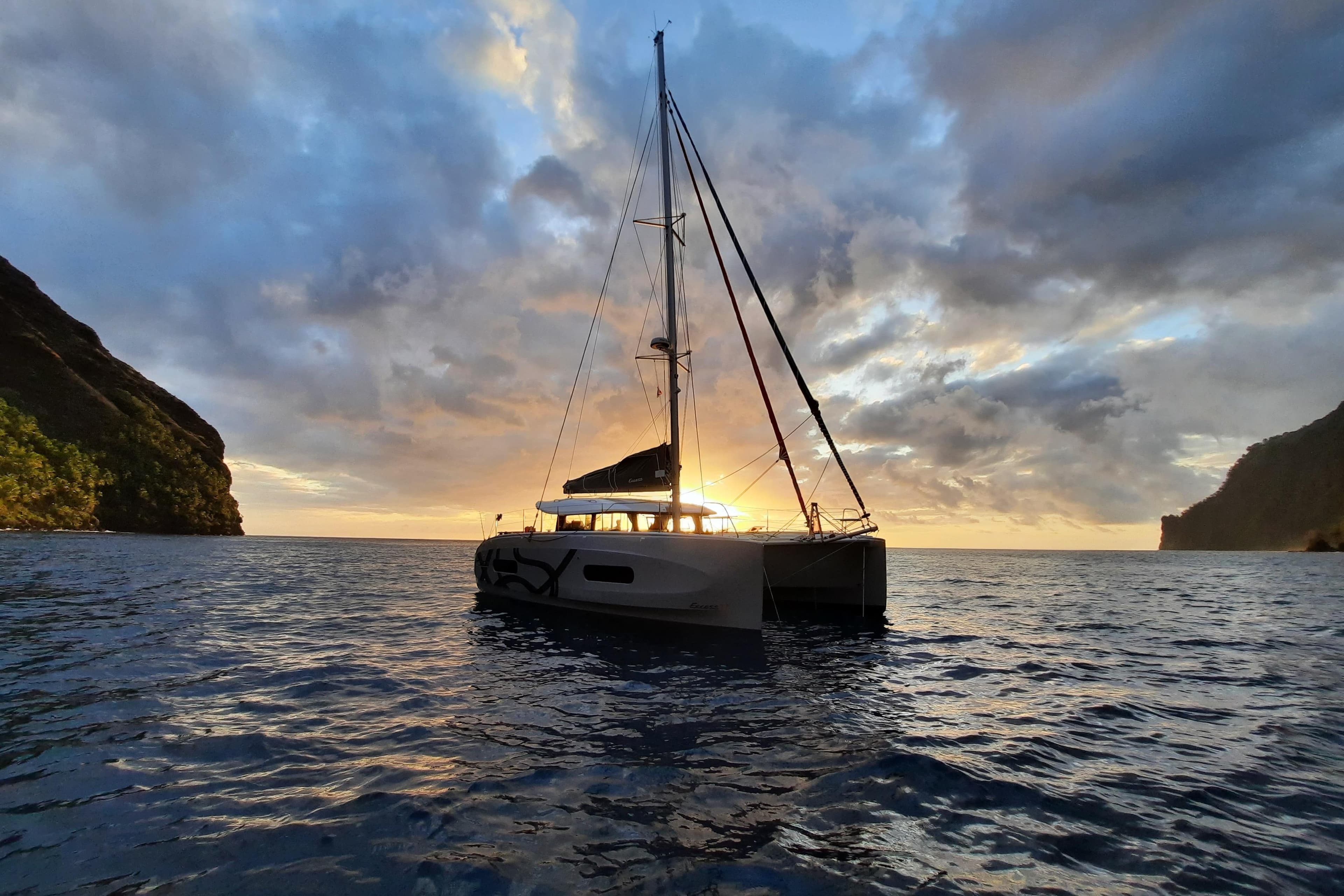 Sailing catamaran Excess 11, 2026 model, at sunset between cliffs on the ocean.
