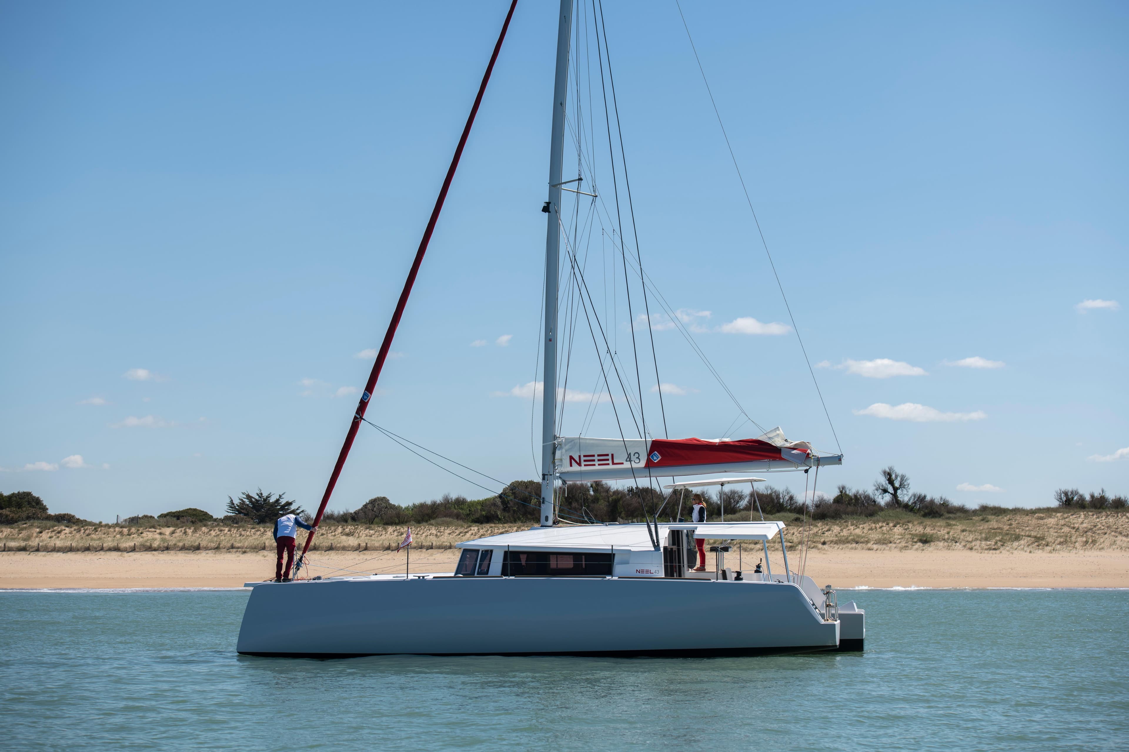 Sailing trimaran NEEL 43, 2025 model, anchored near a sandy beach under a clear blue sky.