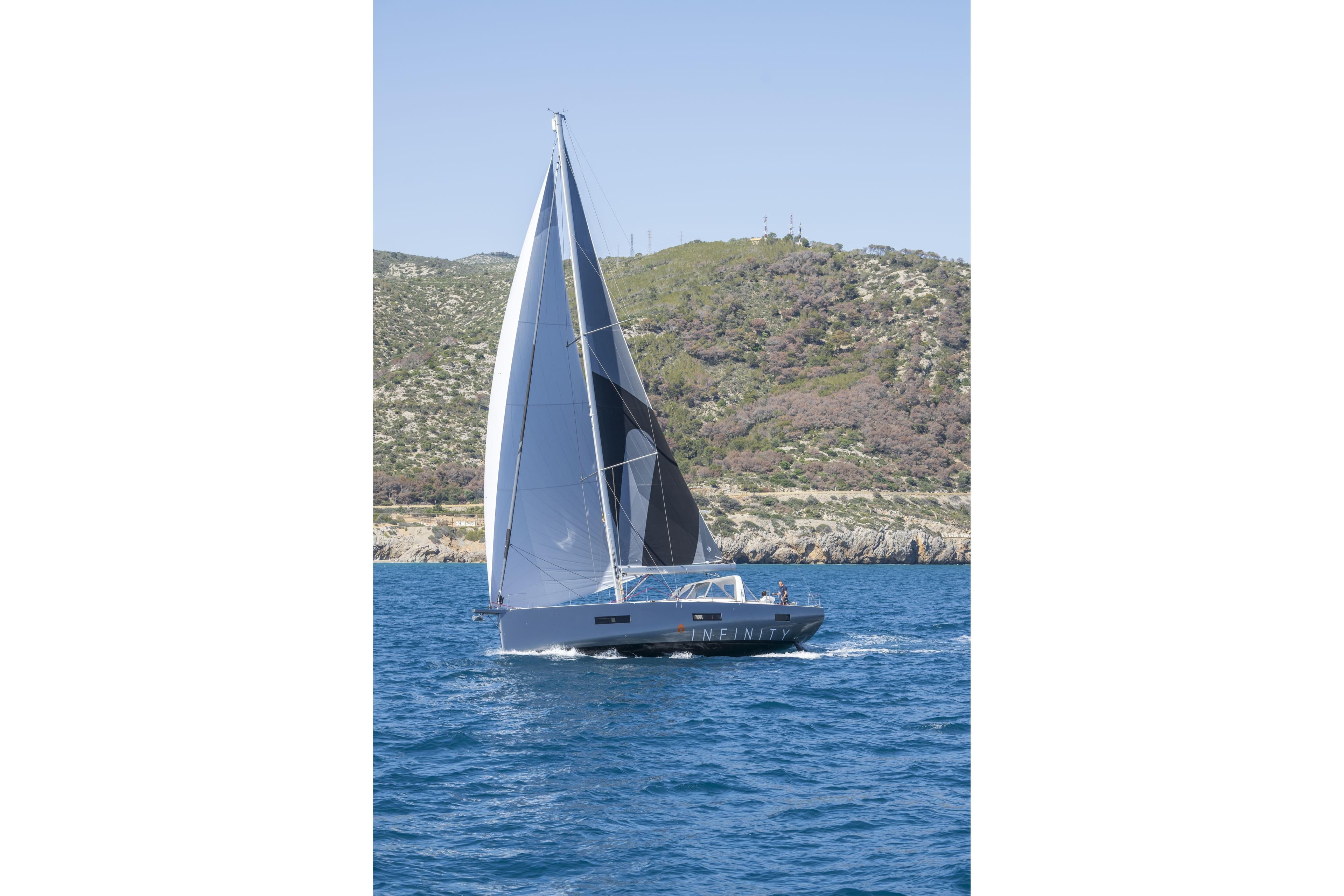 2023 Beneteau Oceanis Yacht 60 sailing near rocky coastline under clear blue sky.