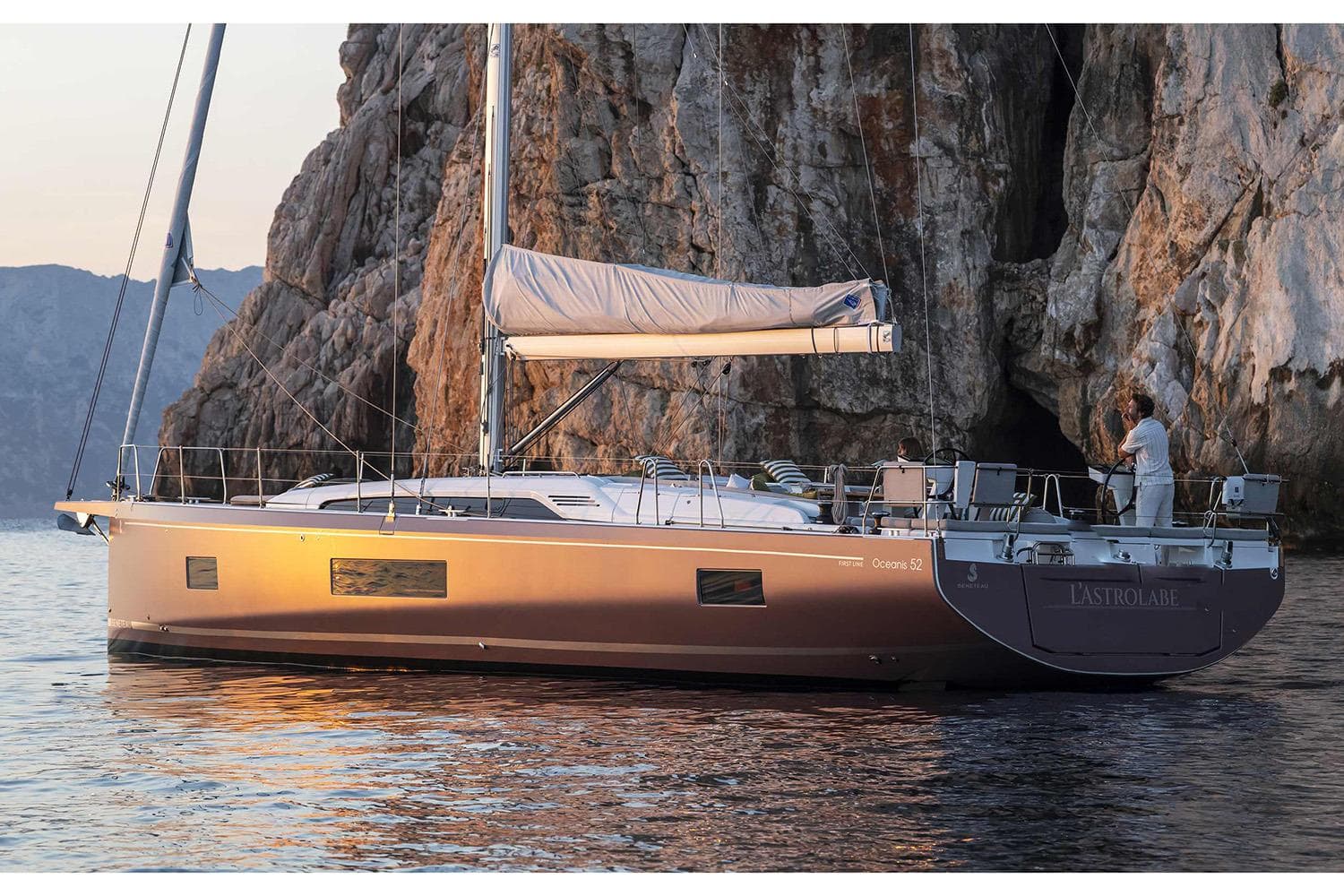 Manufacturer Provided Image: 2026 Beneteau Oceanis 52 yacht sailing near rocky cliffs at sunset.