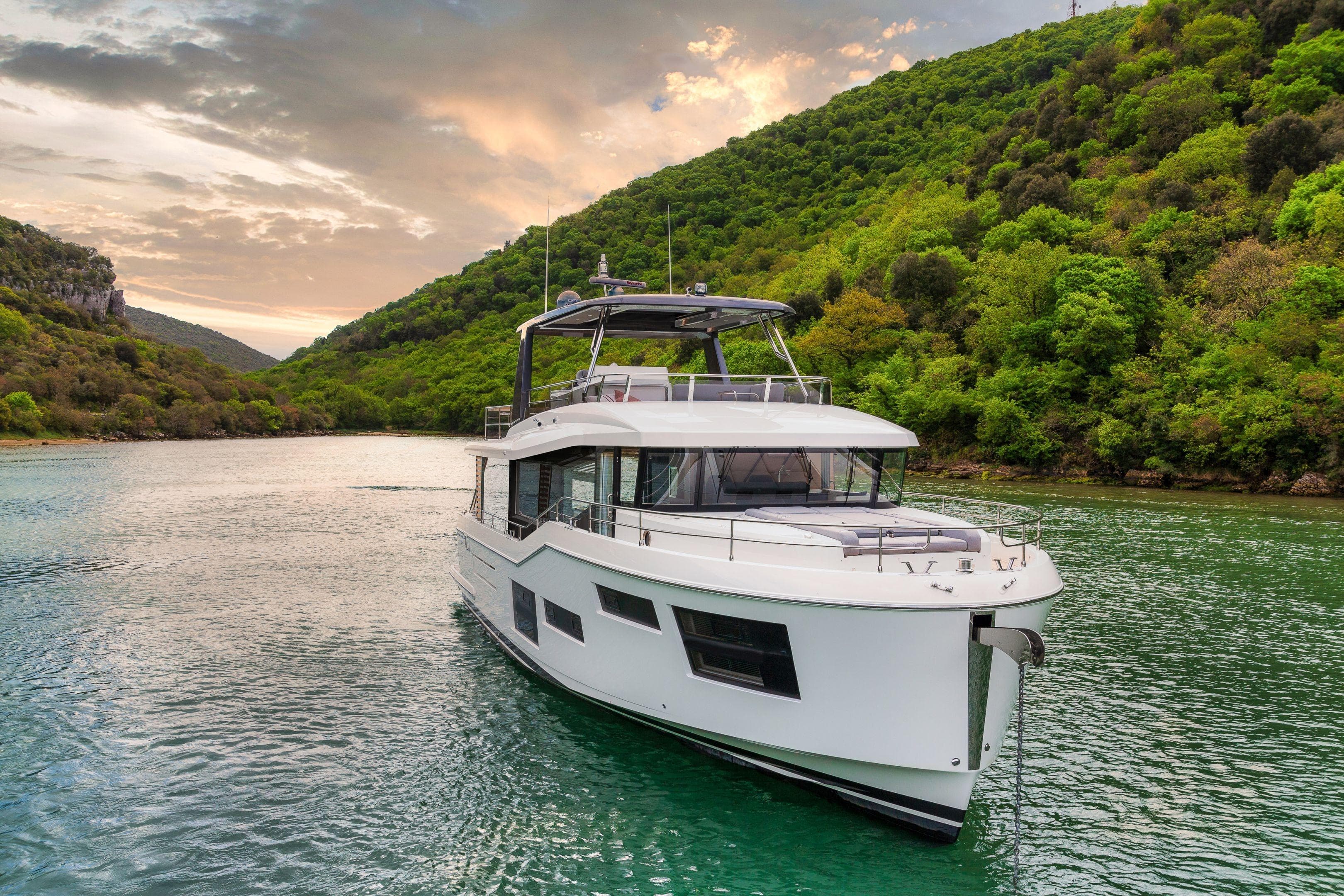 2025 Beneteau Grand Trawler 63 cruising on a scenic river with lush green hills.