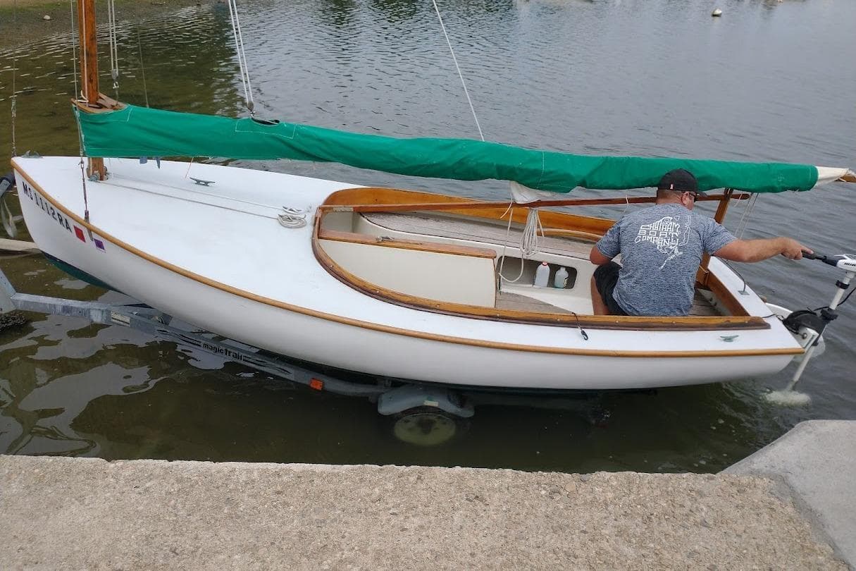 1997 COMPASS CLASSIC Catboat with green sail cover, docked on a trailer by the water.