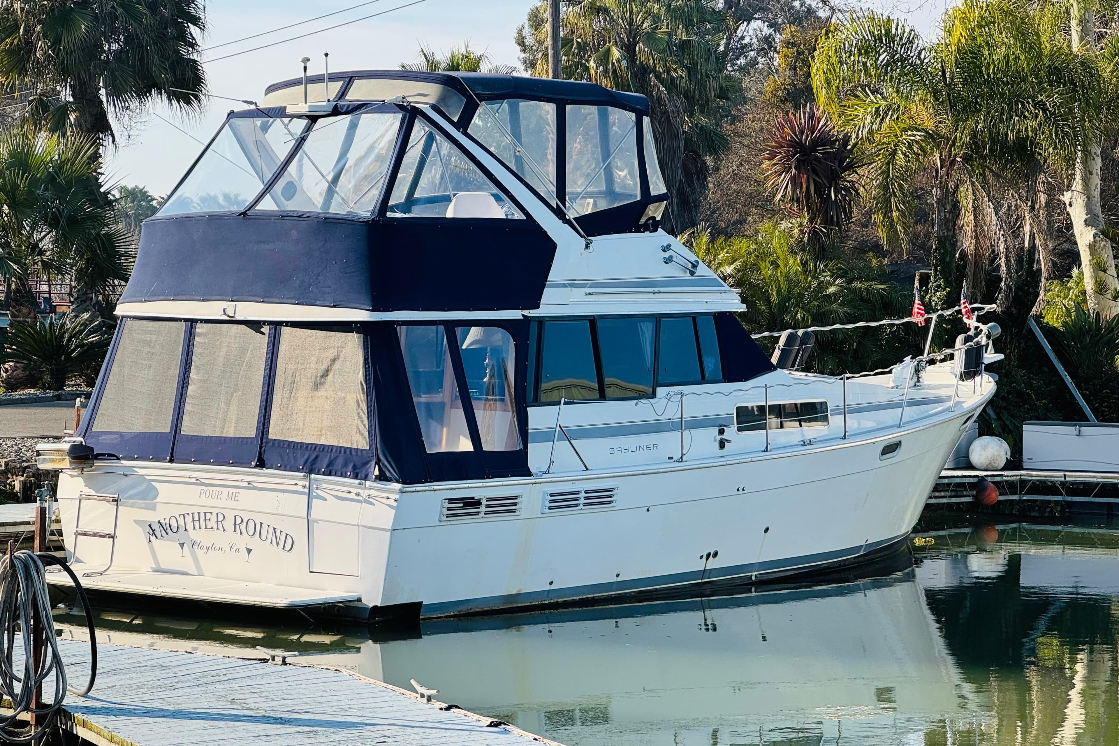1990 Bayliner 3888 Motoryacht docked, featuring blue canopy and lush greenery background.
