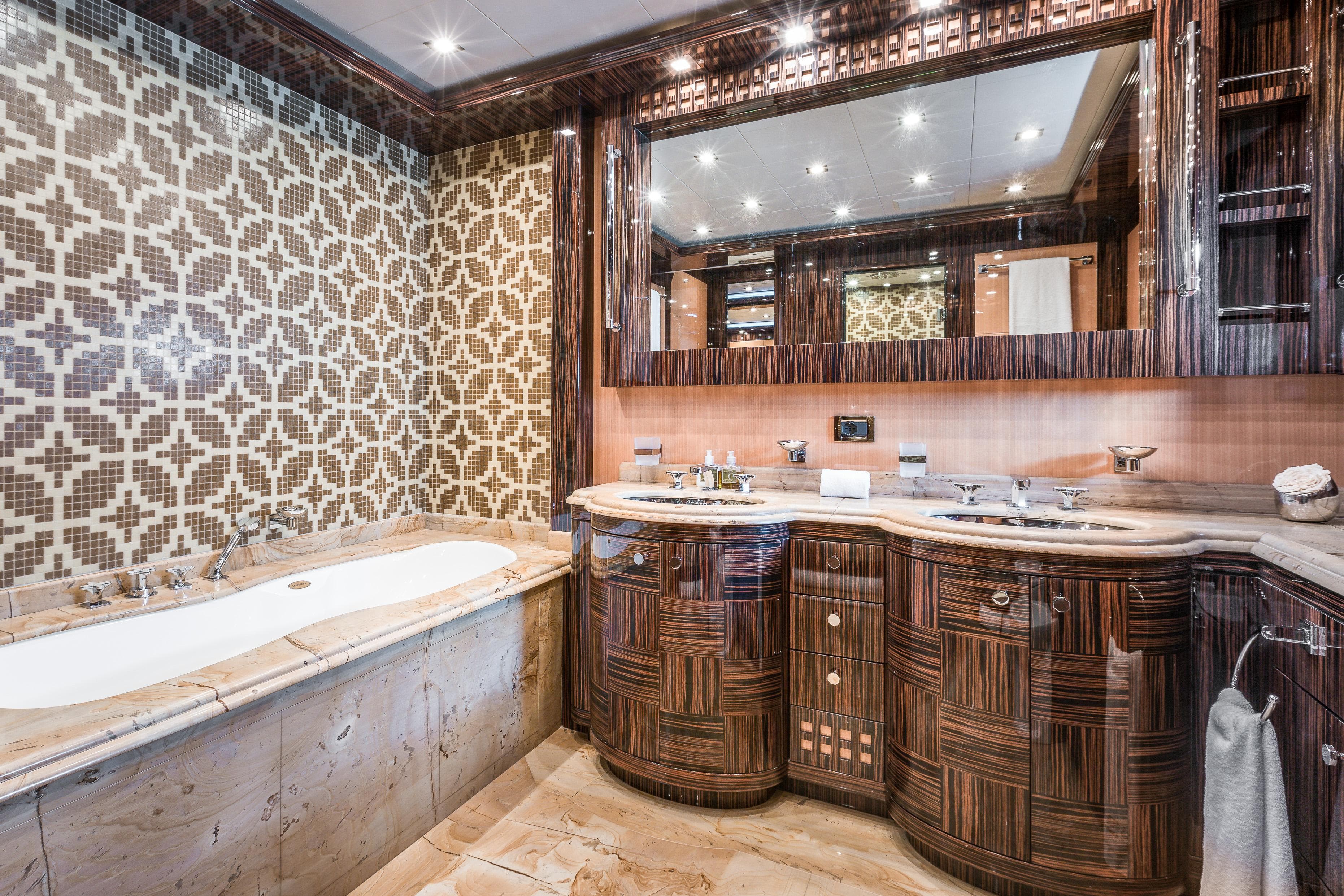 Luxurious bathroom with ornate wood cabinetry, patterned tile wall, and marble bathtub in 2011 Mangusta 165 yacht.