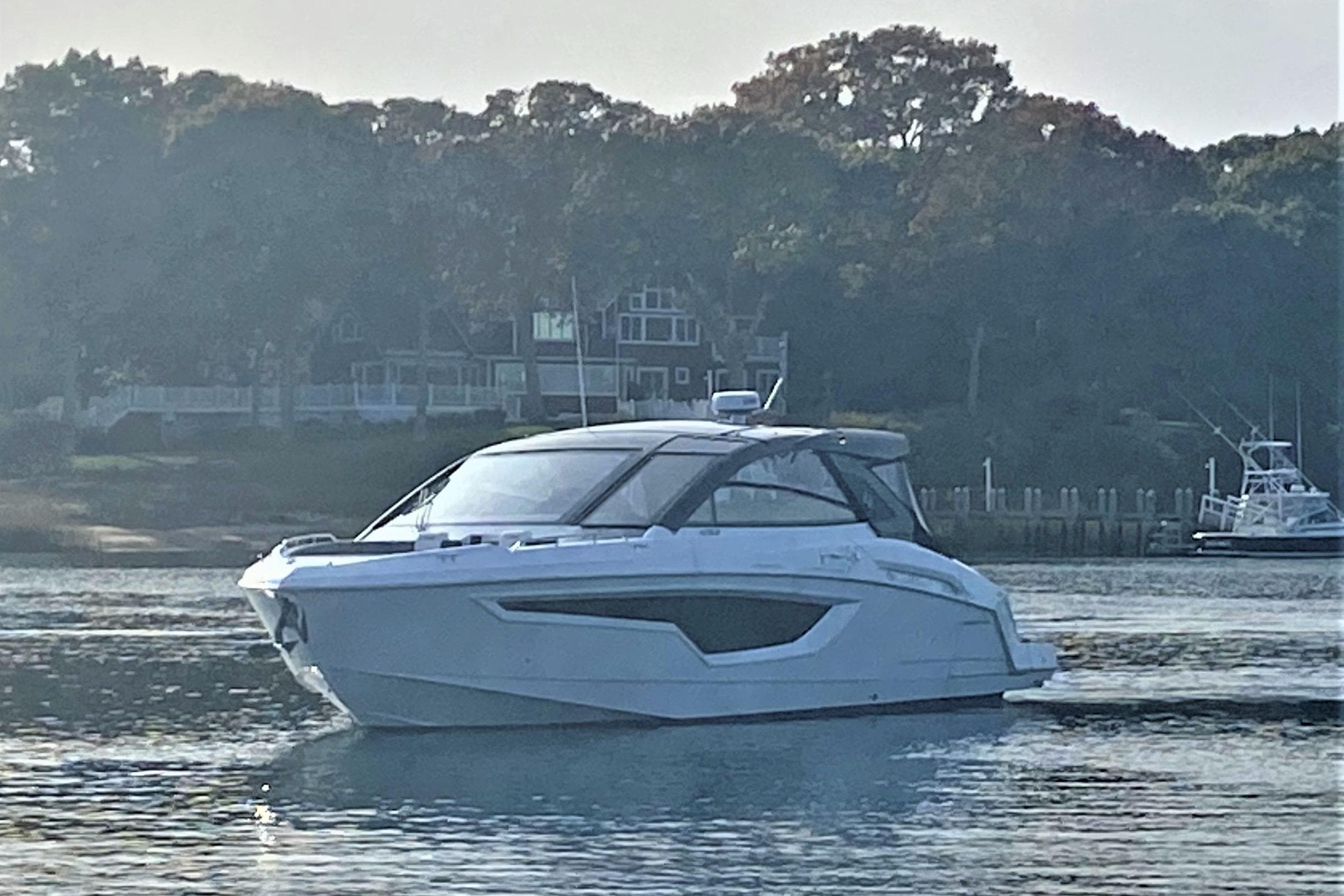 2026 Cruisers Yachts 42 GLS OB on calm water near a wooded shoreline.