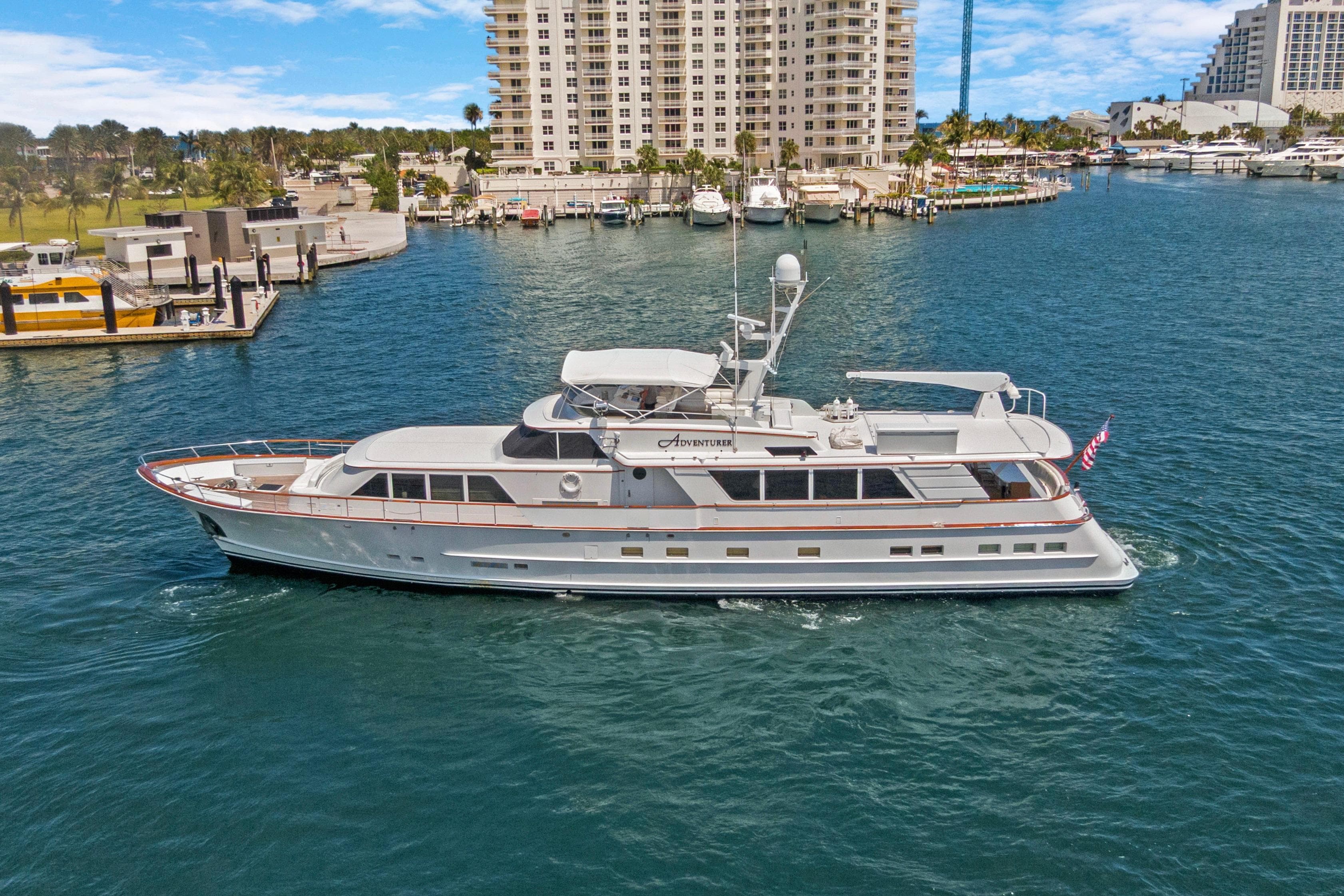 Luxury yacht "Adventurer" cruising near marina, 1985 Burger model, clear skies, waterfront buildings.