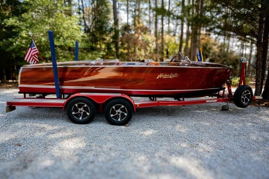1941 Hacker-Craft Deluxe boat on red trailer, surrounded by trees.