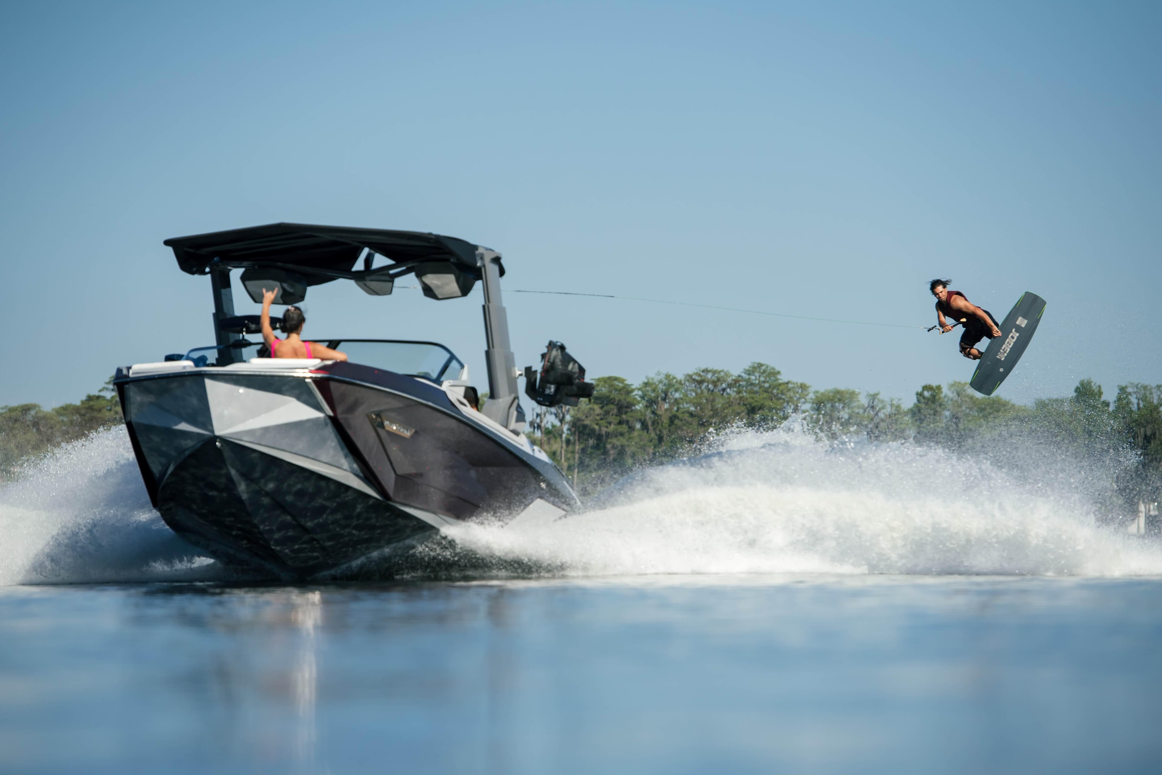 Wakeboarder jumps behind 2026 Nautique G23 PARAGON boat on a sunny day.