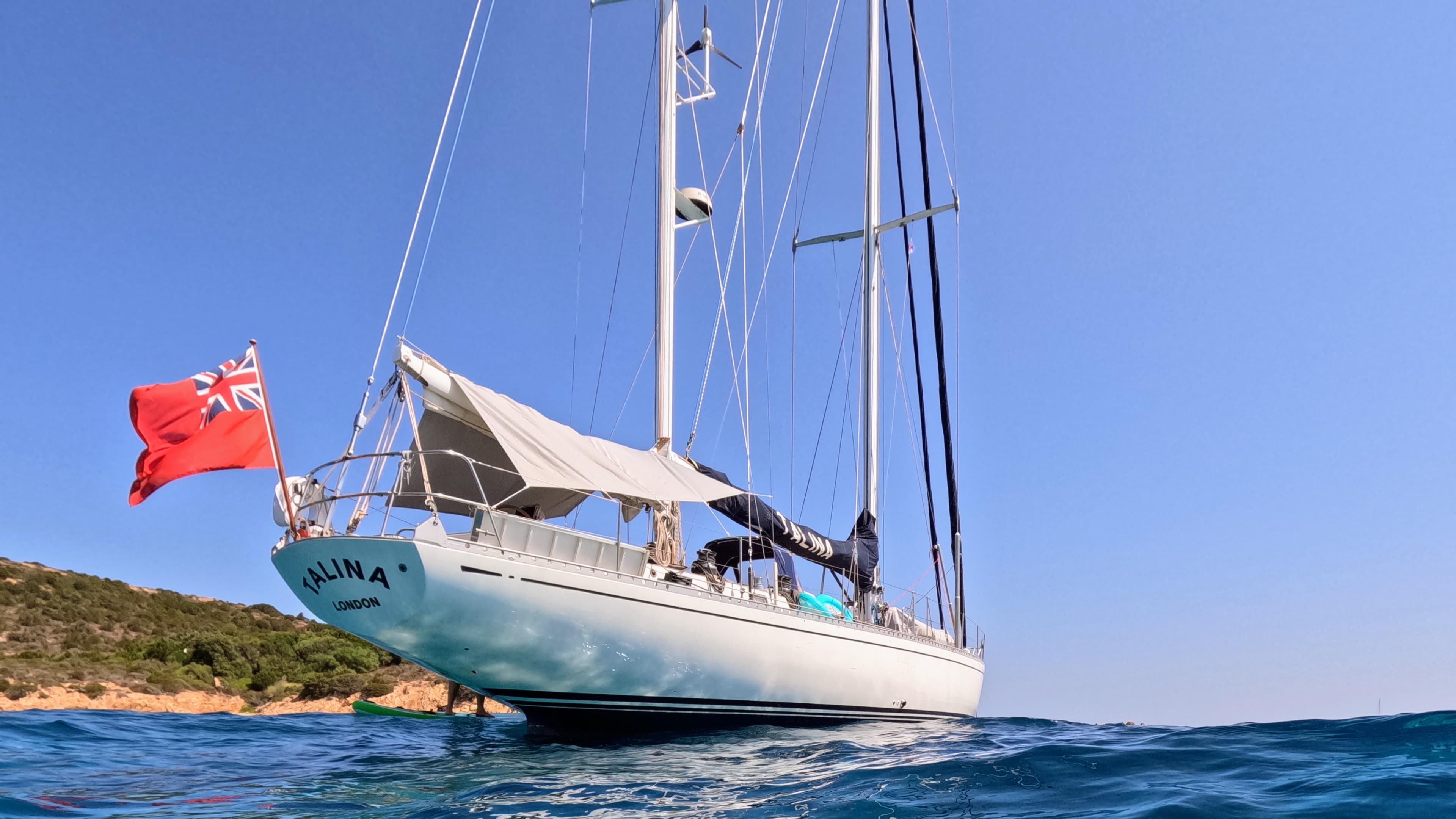Sailing yacht Nautor Swan 65 (1975) with red flag, anchored in clear blue waters.