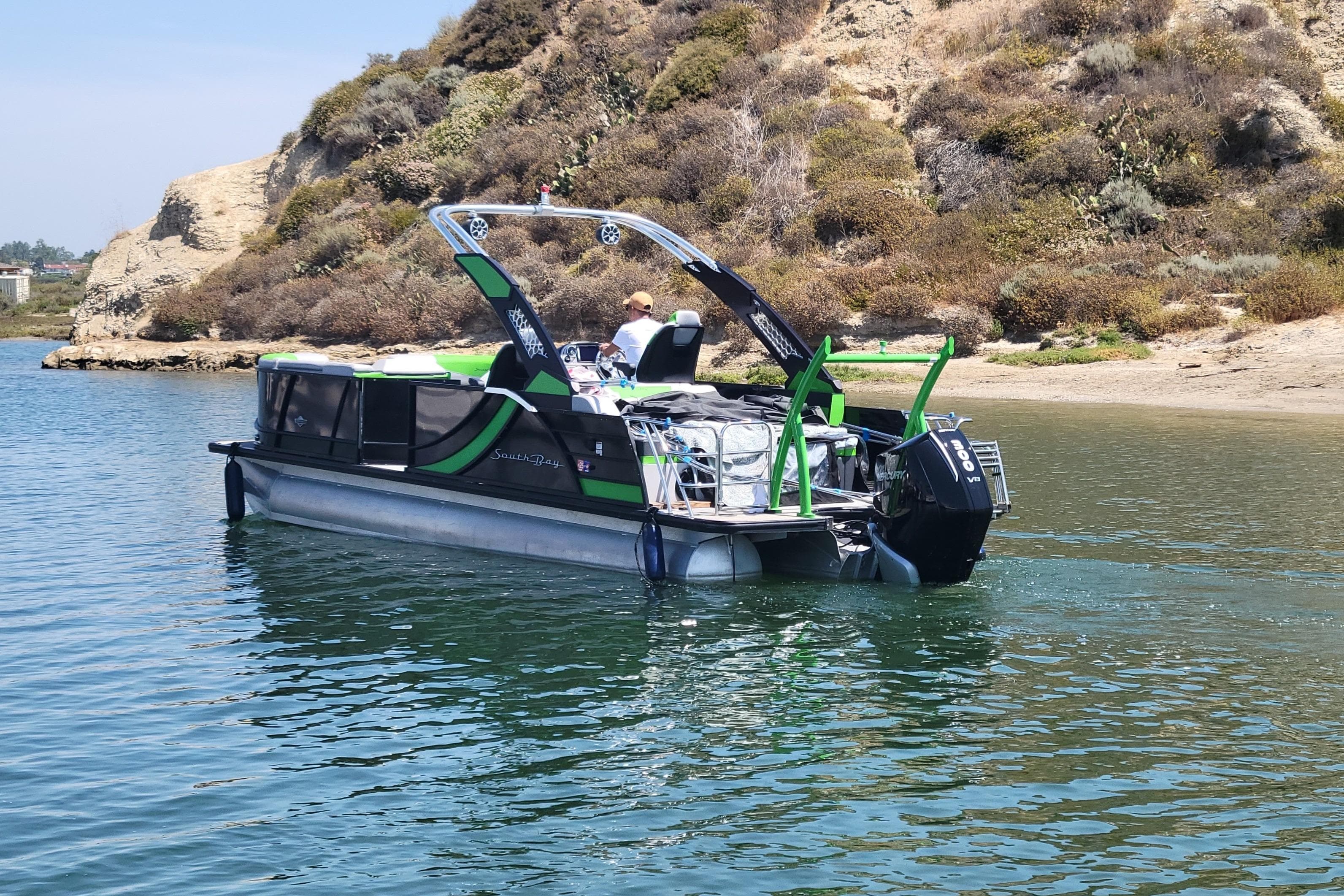 2023 South Bay 525UL AR pontoon boat on calm water near rocky shoreline.