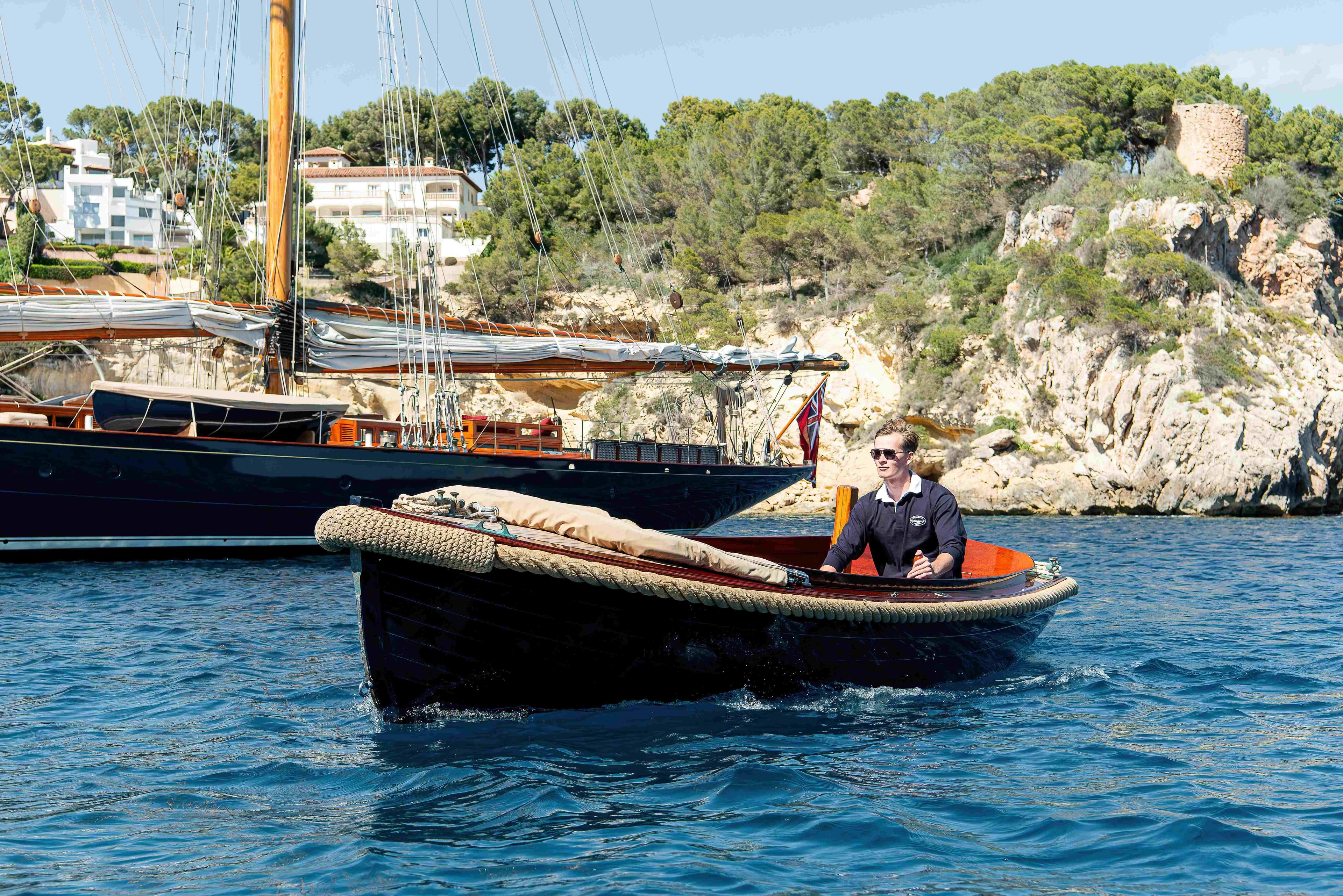 Person in a small boat near a 1915 Herreshoff Performance Schooner, coastal background.