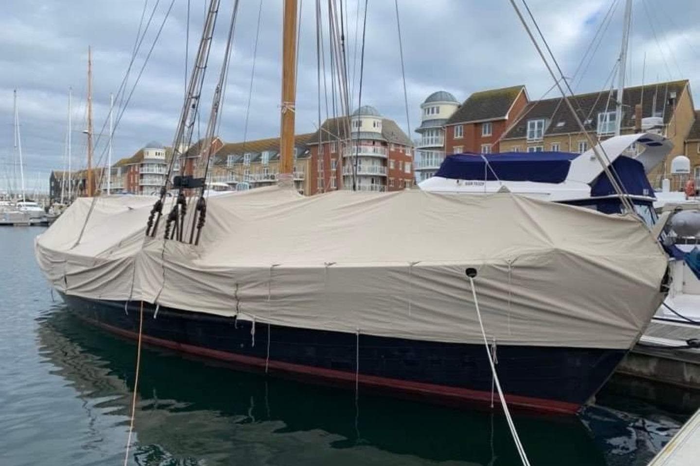 Classic 1899 Bristol Pilot Cutter covered at marina, with waterfront buildings in background.