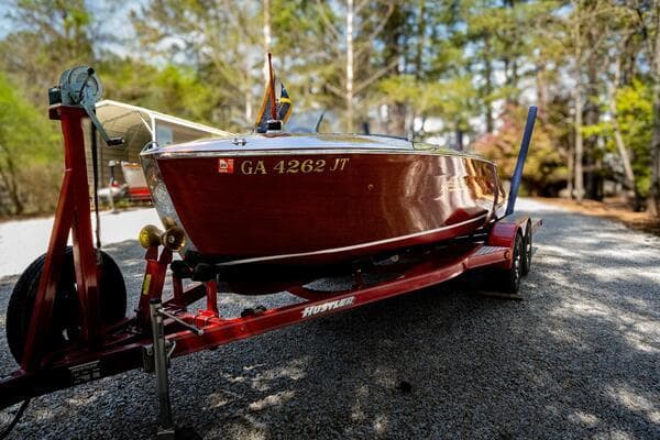 1941 Hacker-Craft Deluxe boat on red trailer, surrounded by trees.