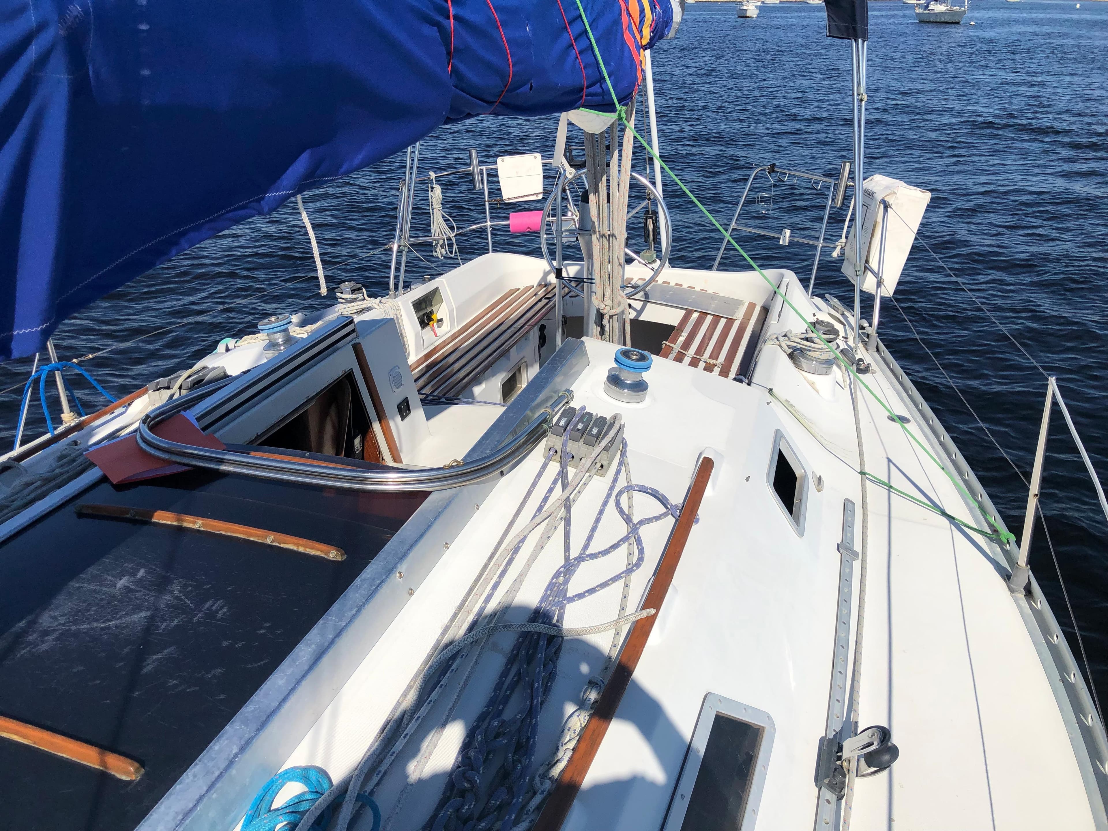 1985 Beneteau First 345 sailboat deck with blue sail, moored on calm water.