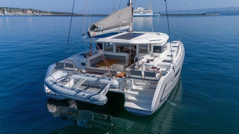 2022 Excess 12 catamaran on calm water with dinghy attached, under clear blue sky.