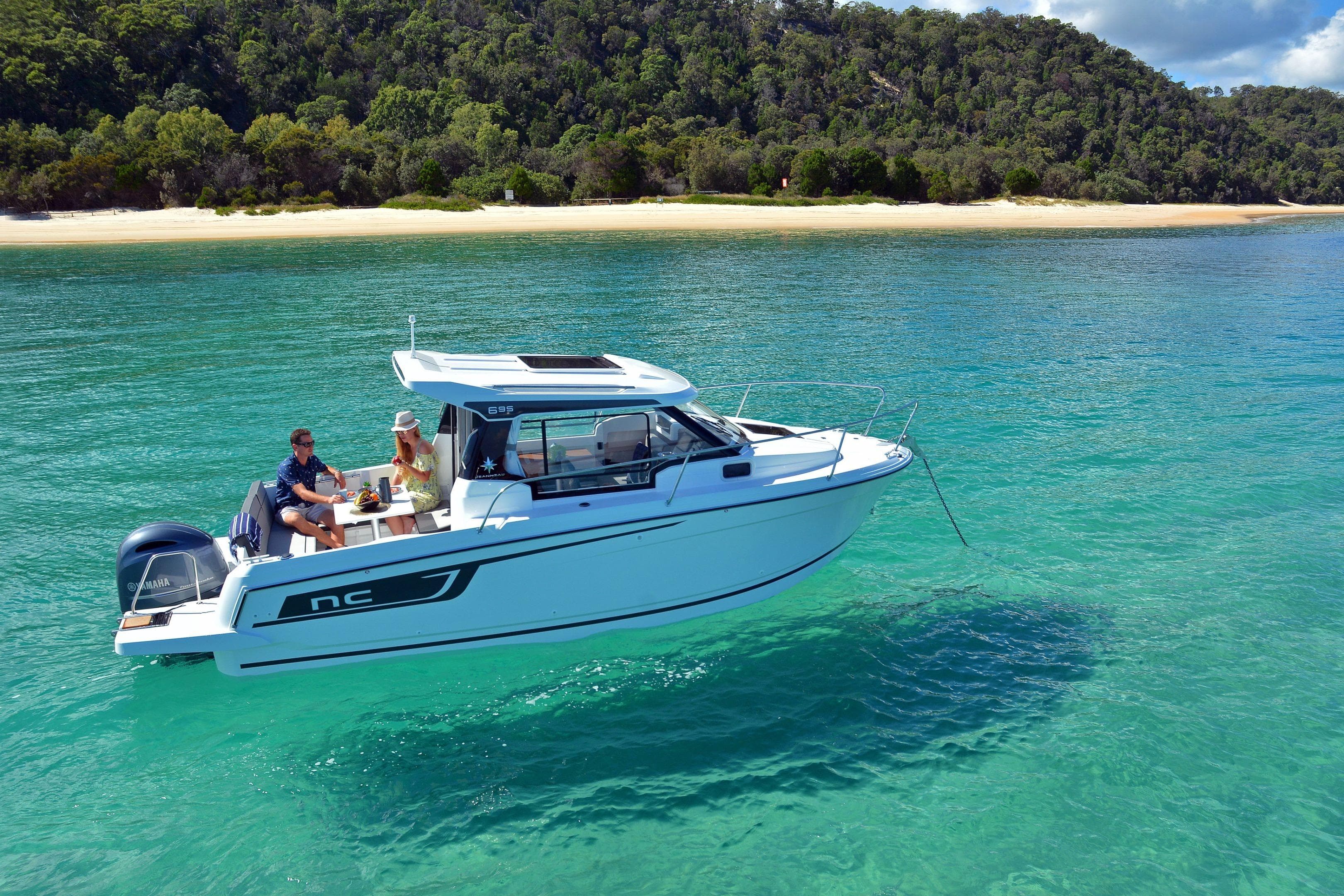 2026 Jeanneau NC 695 boat cruising on turquoise waters near a lush, sandy beach.