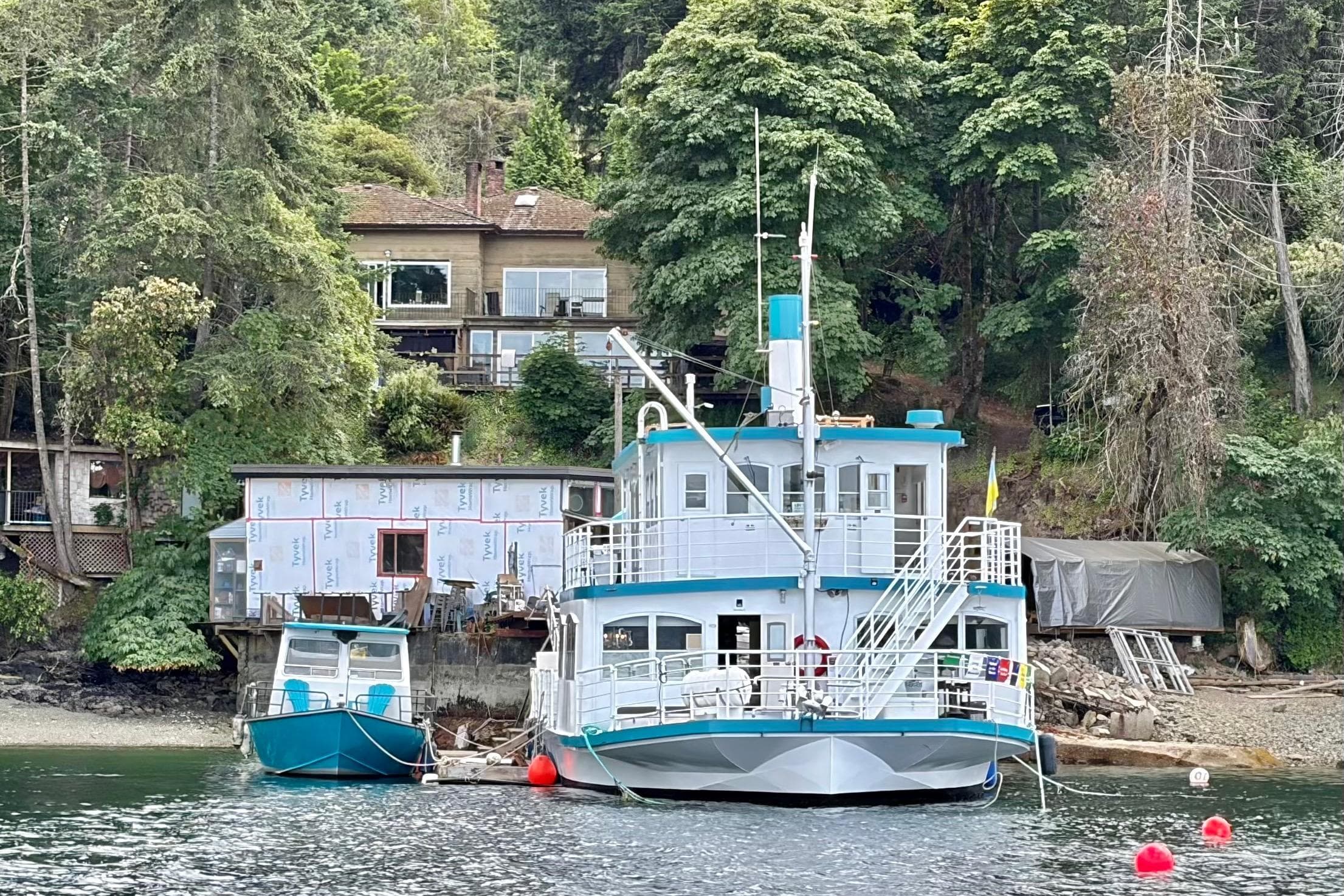1988 Titan houseboat conversion docked by a forested shoreline with nearby buildings.