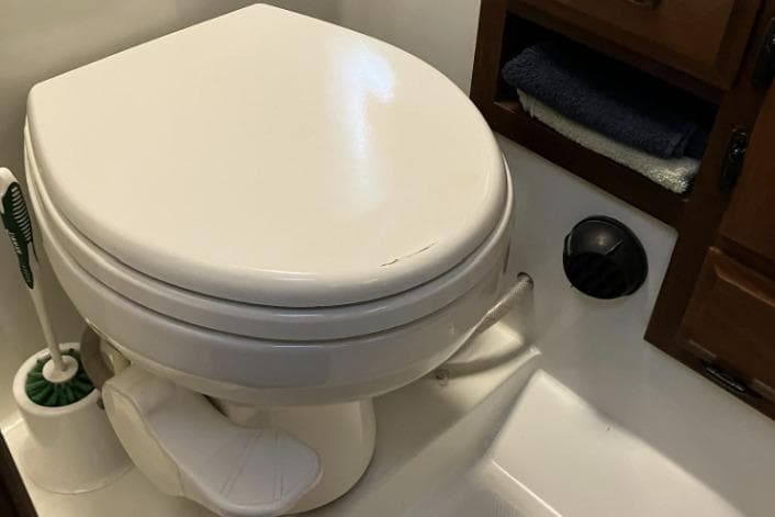 White RV toilet with foot pedal, adjacent storage, and cleaning brush.