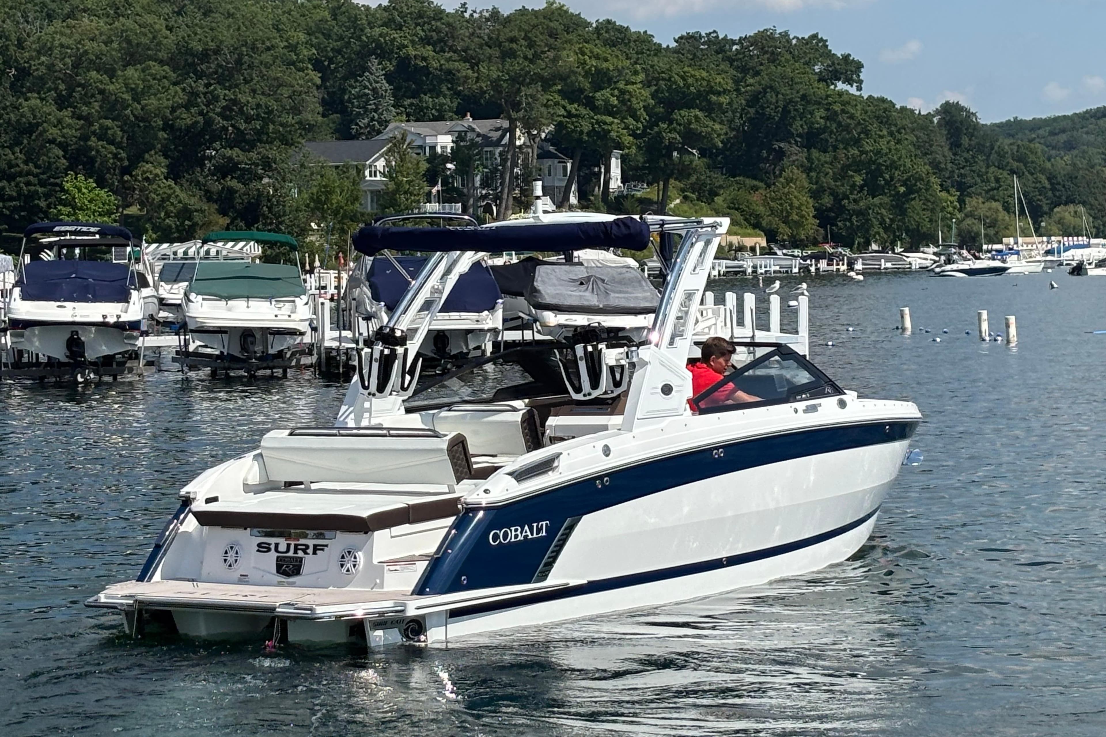 2025 Cobalt R6 Surf boat cruising on a scenic lake near a marina.