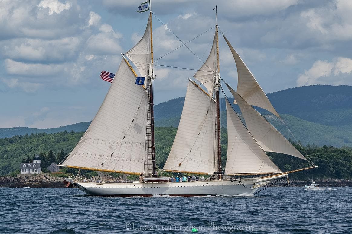 Classic 1962 Coasting Schooner sailing near a scenic coastline with lush greenery and mountains.