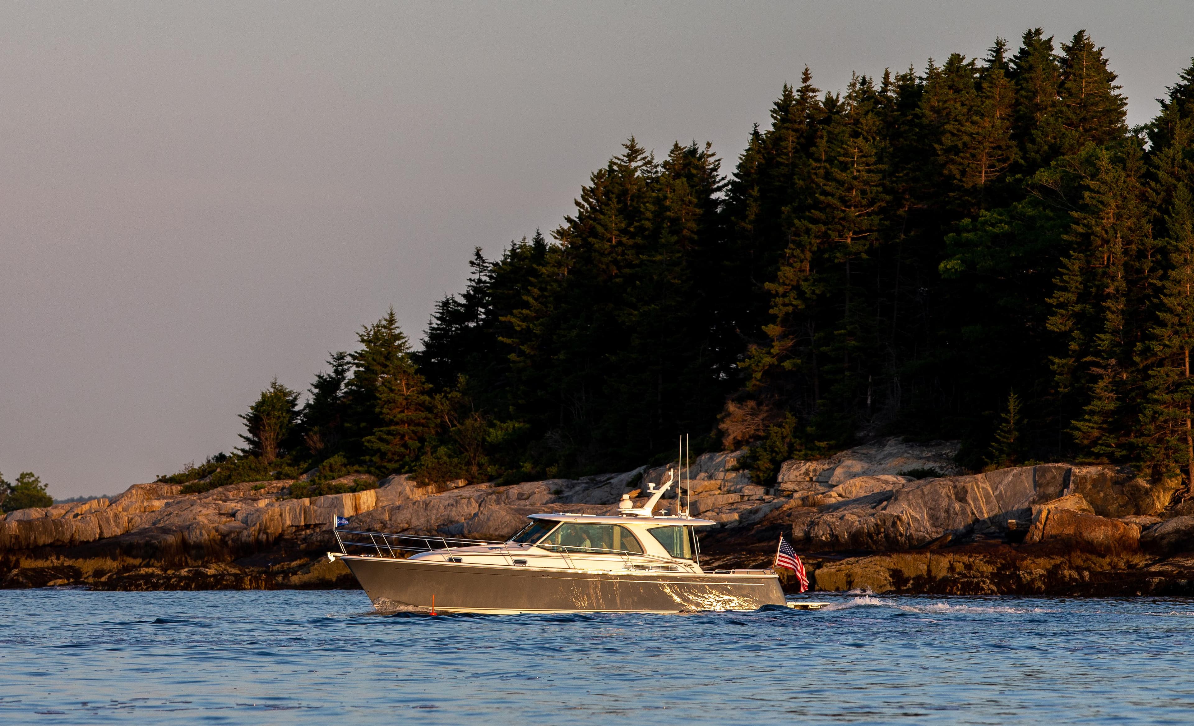 2025 Sabre 43 Salon Express yacht cruising near a forested rocky shoreline.