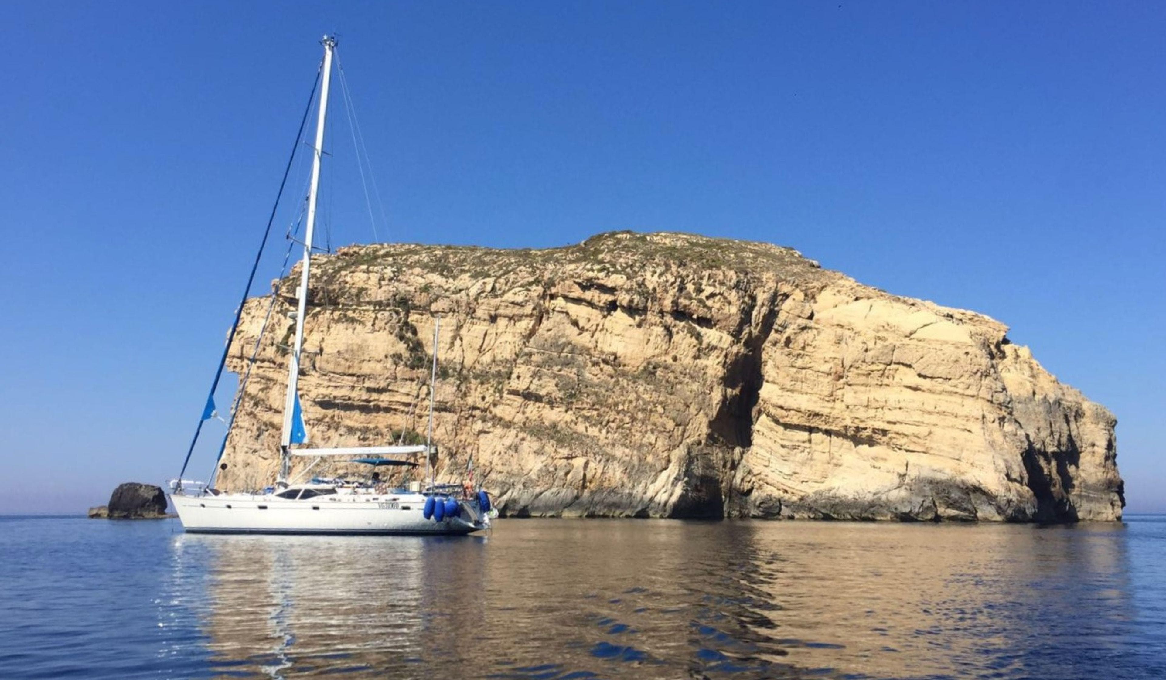 Sailing yacht Oyster 56 (2000) near a rocky cliff under a clear blue sky.