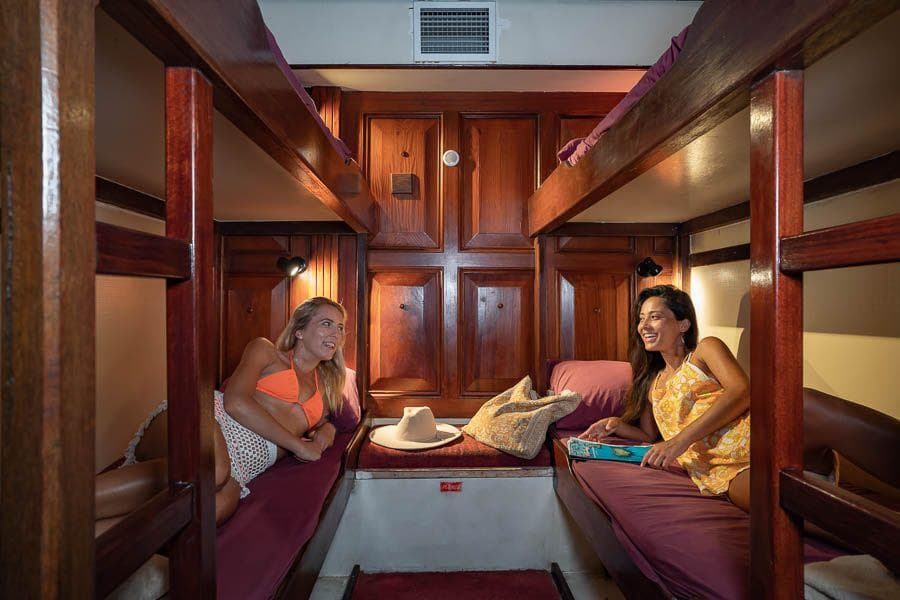Two women relaxing in a cozy wooden cabin with bunk beds on a vintage sailing vessel.