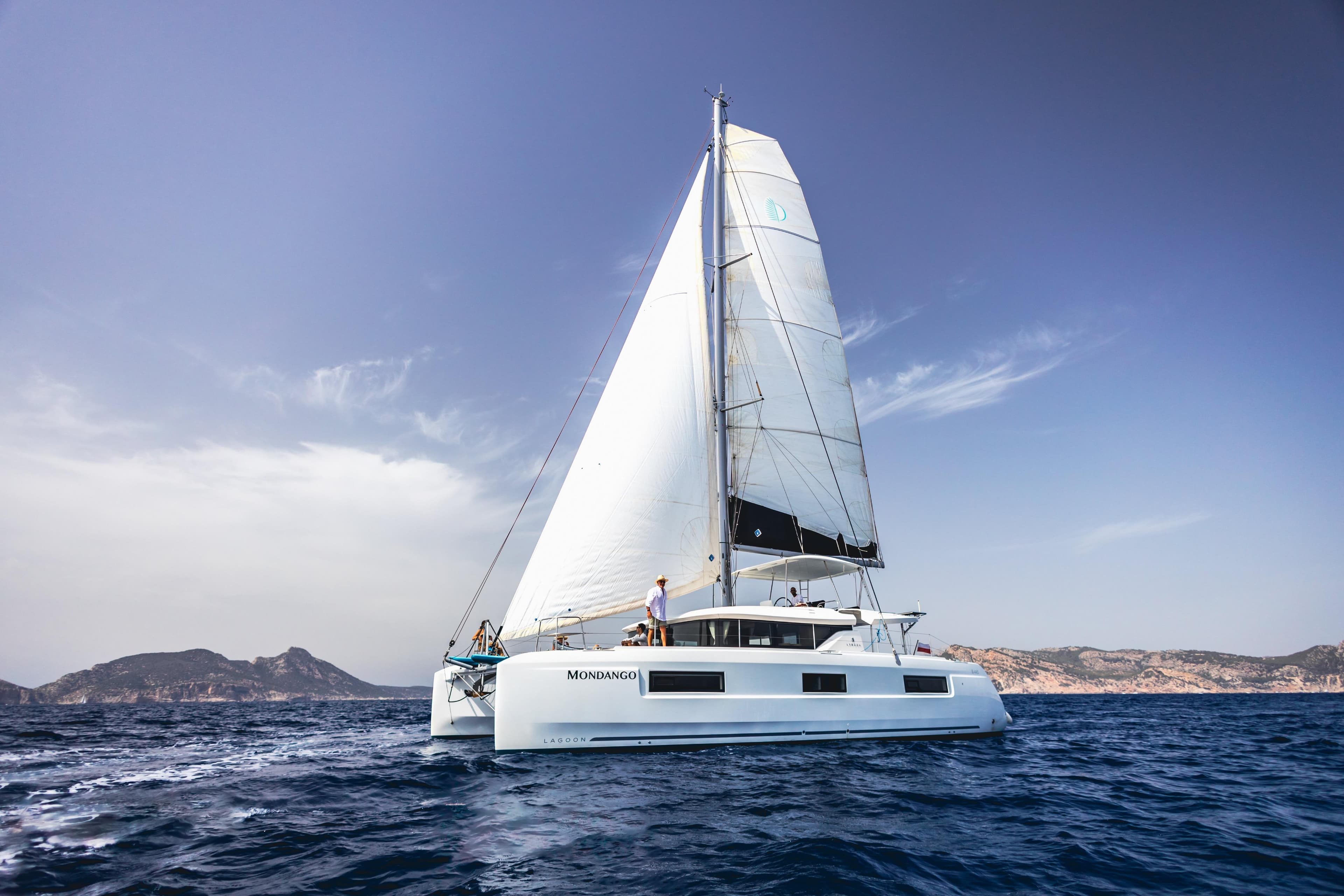 2020 Lagoon 46 catamaran sailing on open sea with mountainous backdrop.