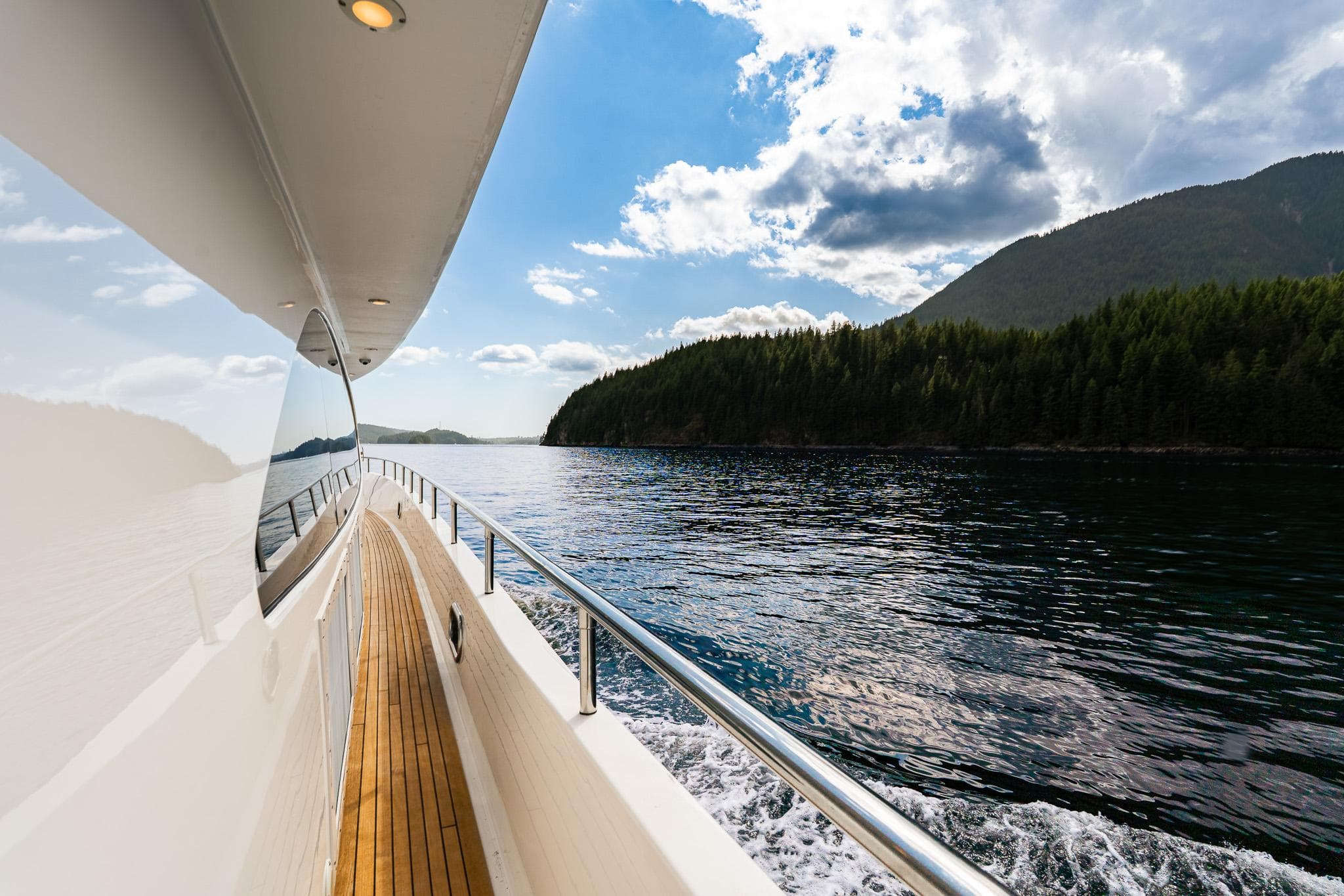 Luxurious Mystica Skylounge yacht cruising near forested coastline under a partly cloudy sky.