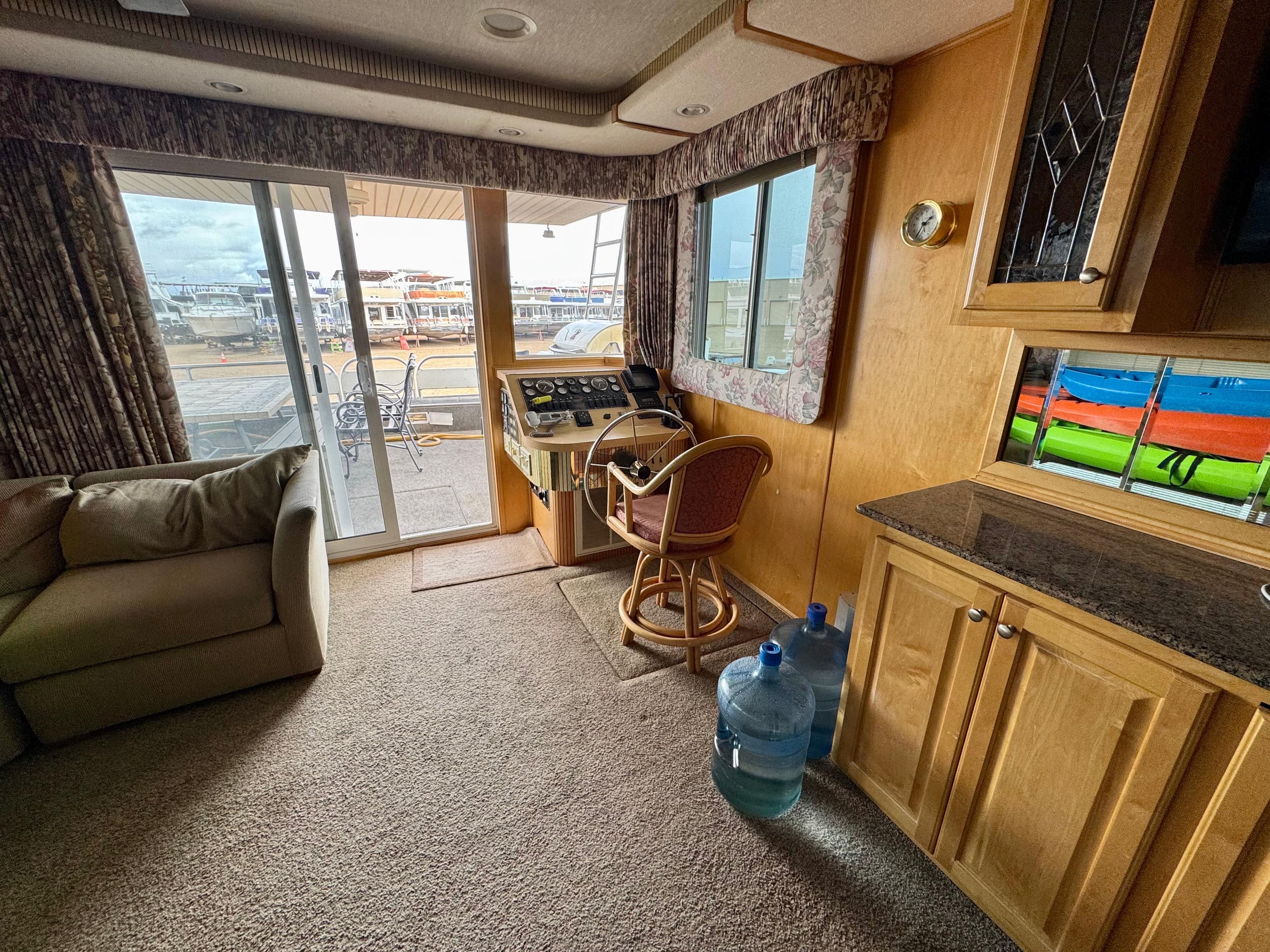 Cozy houseboat interior with helm, sofa, and water jugs, featuring large windows and sliding doors.