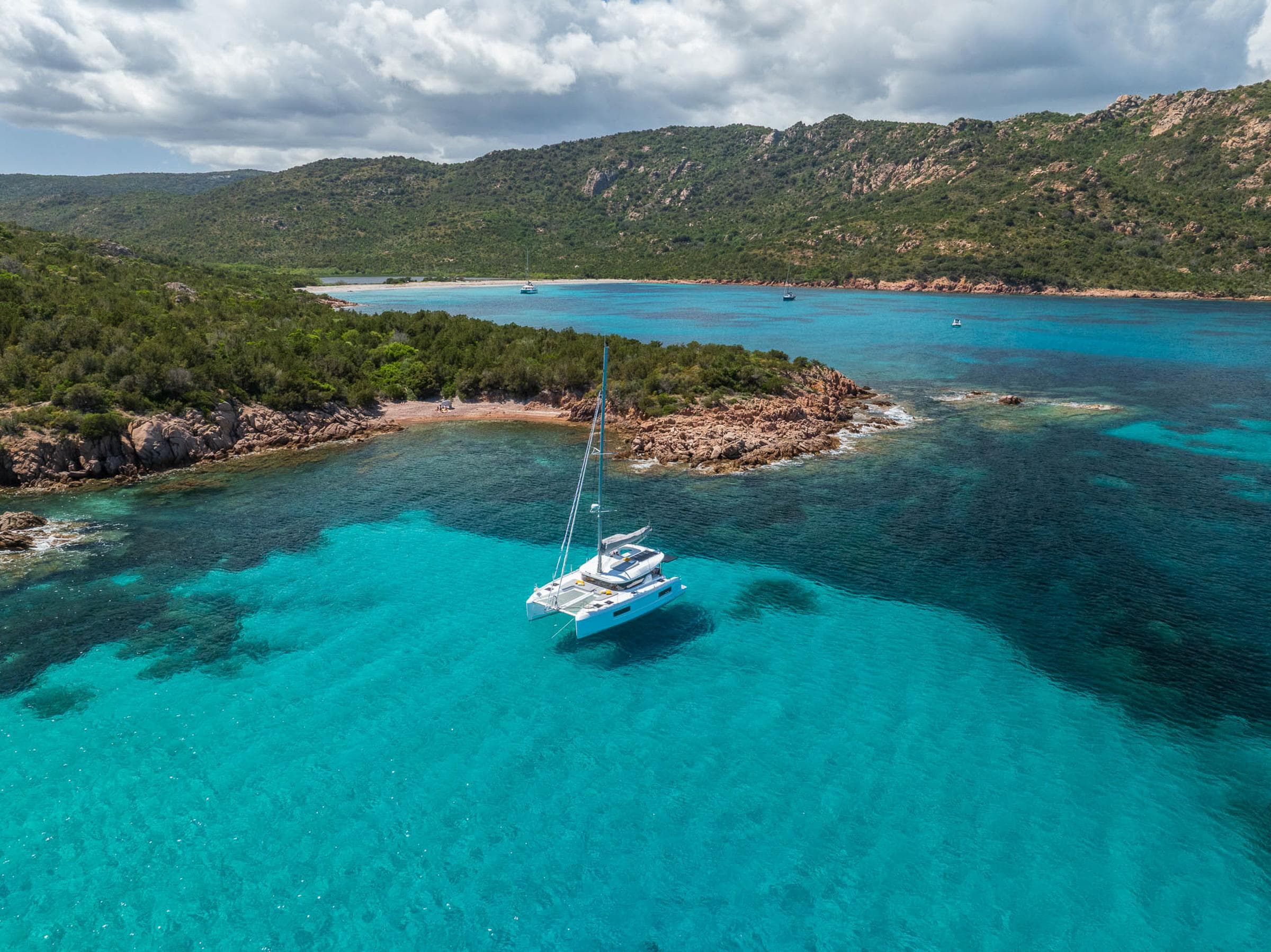 2025 Lagoon 38 catamaran sailing in clear turquoise waters near a rocky coastline.