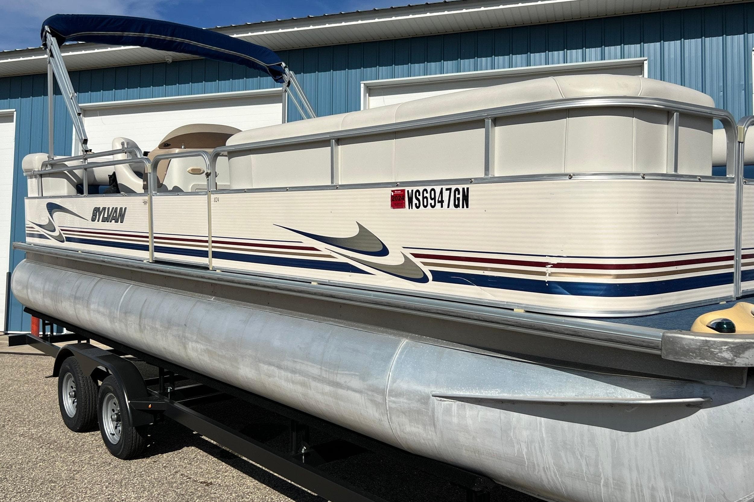 2003 Sylvan 824 Mirage Cruise pontoon boat on trailer, parked near blue building.