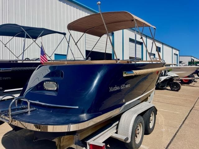 2023 Maxima 620 Retro boat on trailer, blue hull, tan canopy, parked outside.