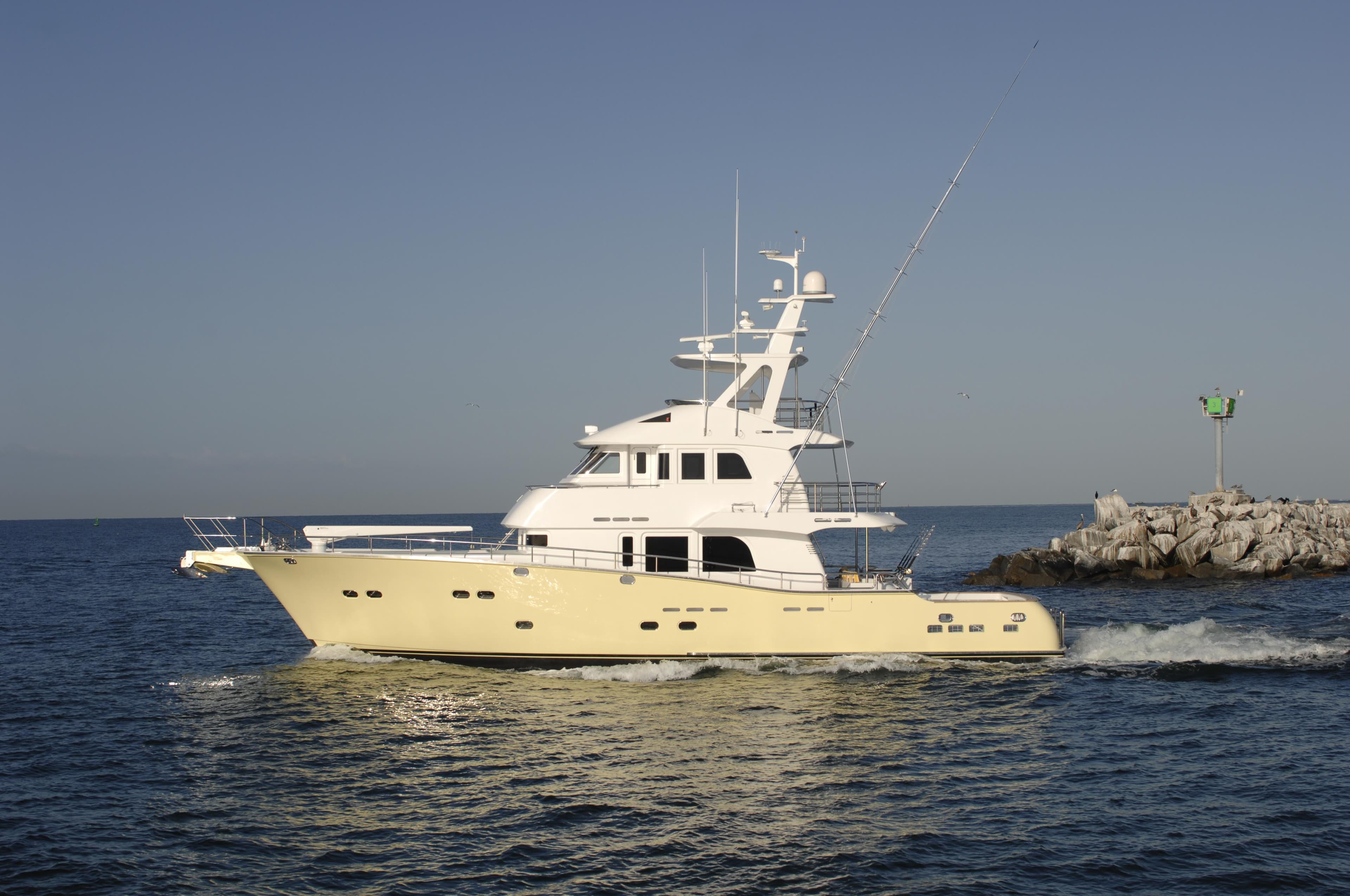 2025 Nordhavn 75 Expedition Yachtfisher cruising near rocky shoreline under clear sky.