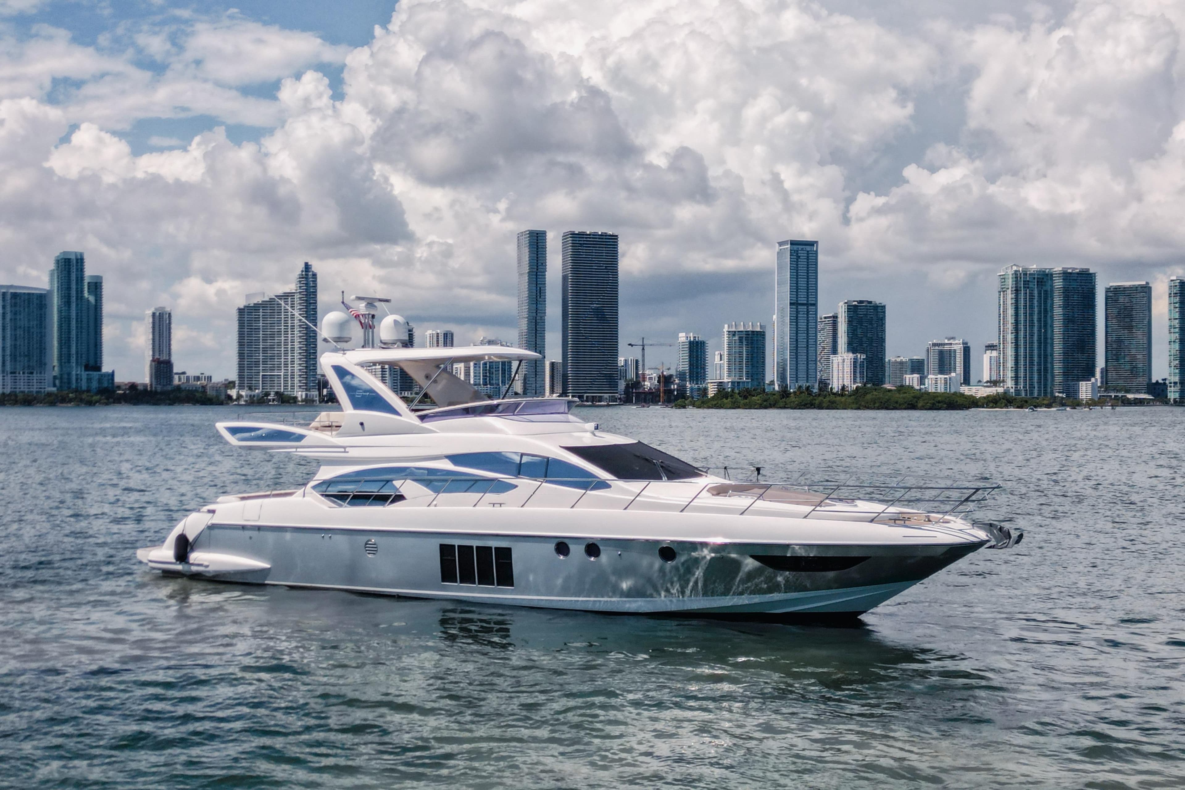 2016 Azimut 64 Flybridge yacht cruising near city skyline under cloudy sky.