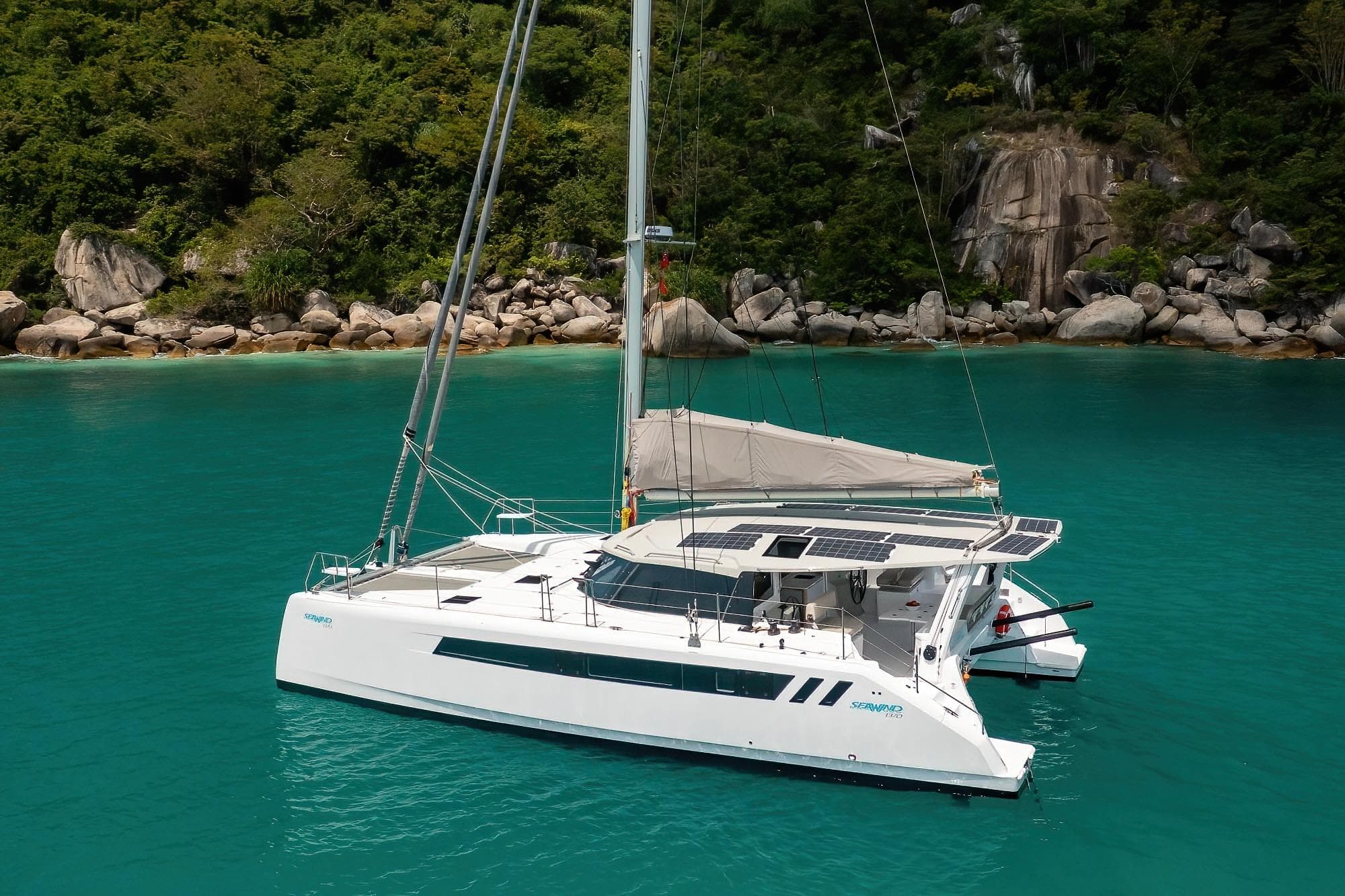 2026 Seawind 1370 catamaran sailing in turquoise waters near a rocky, forested shoreline.