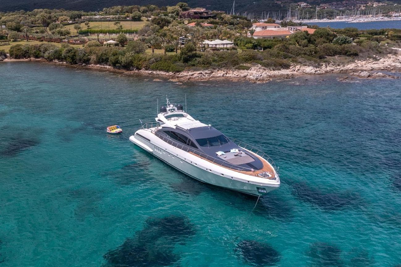 Luxurious 2006 Mangusta 92 yacht anchored in clear turquoise waters near a scenic coastline.
