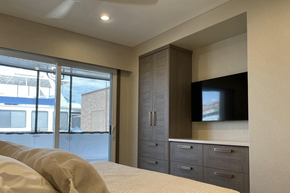 Modern bedroom interior with large window, sleek cabinetry, and wall-mounted TV in Adonia model.
